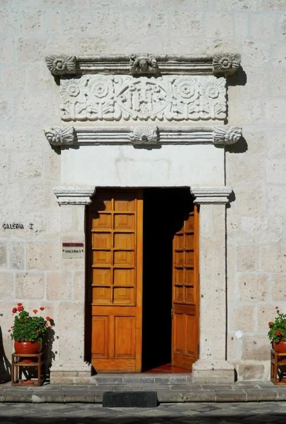 A stone building with a carved decorative lintel above a wooden door, flanked by potted plants with red flowers.