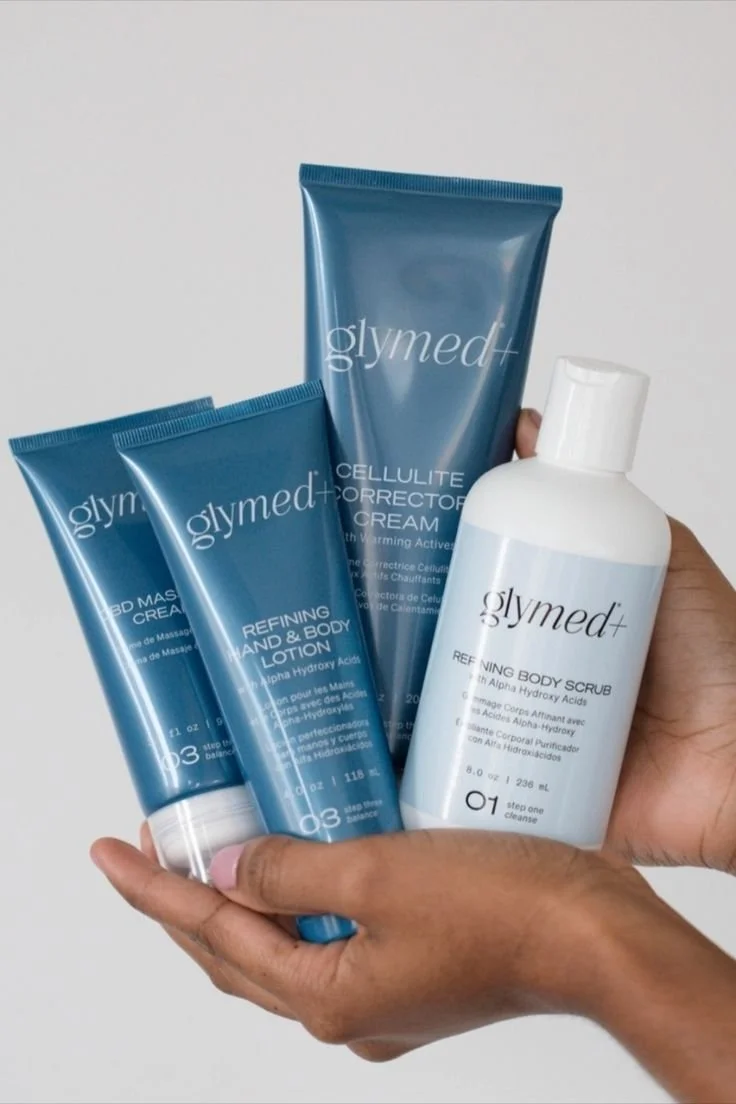 Hand holding skincare products from Glymed+ including a body scrub, hand and body lotion, a cream, and a refining hand and body lotion.