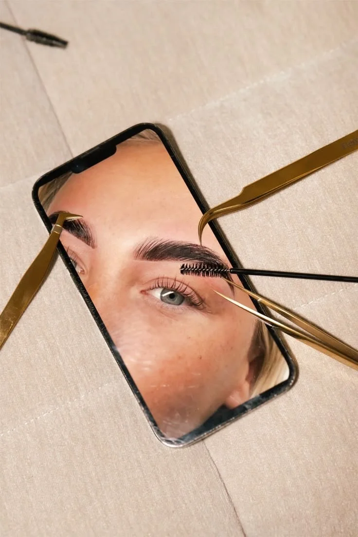 Close-up photo of a smartphone screen reflecting a woman with well-groomed eyebrows and blue eyes, with eyelash extension tools positioned near her eye.