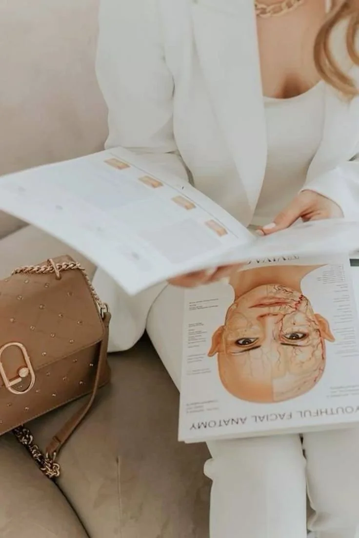 A person wearing a white outfit sitting on a beige couch, reading a magazine about the facial anatomy of a human face. A light brown handbag with a chain strap is placed next to them.