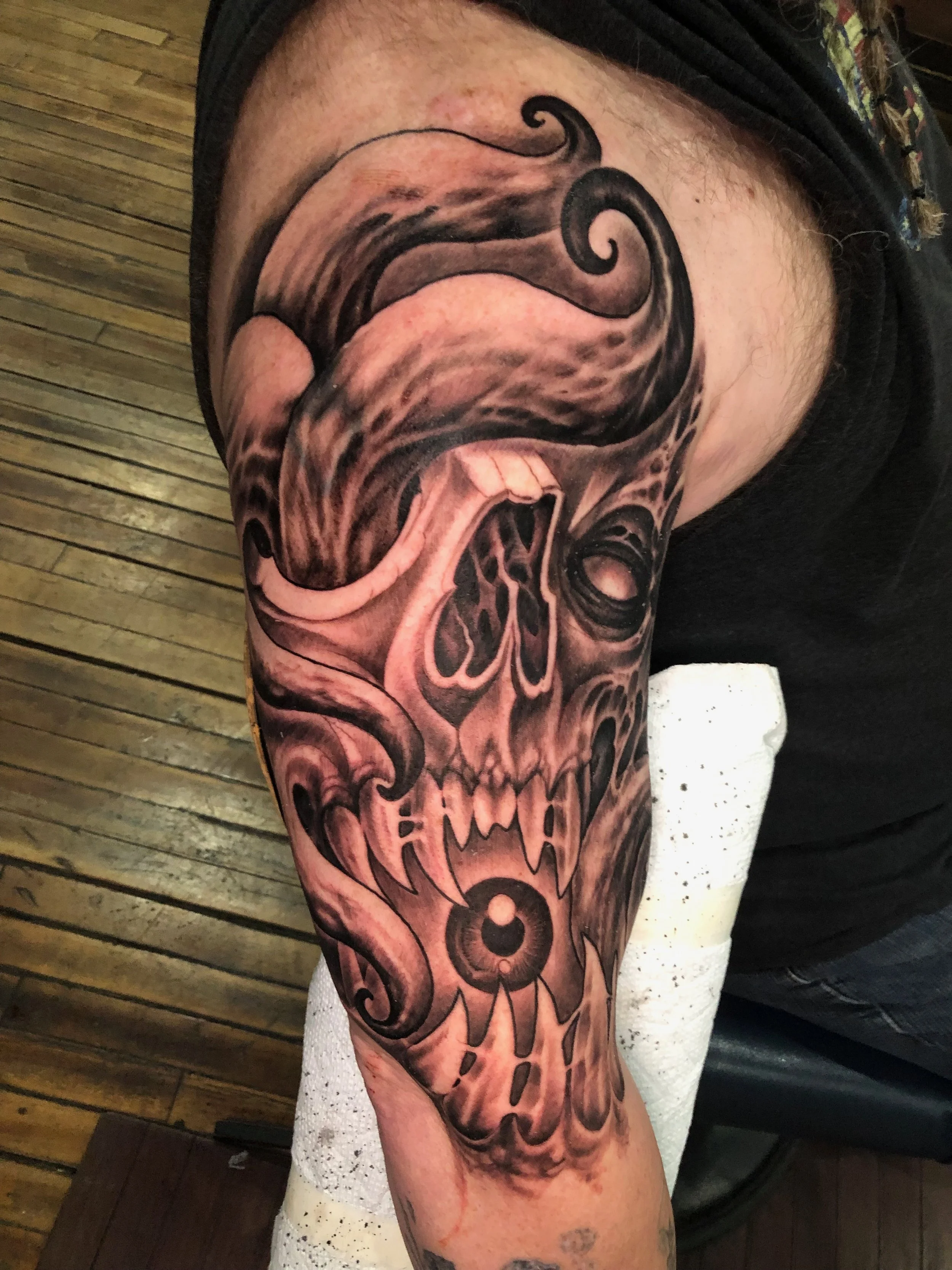 A tattoo of an angry skull with a stylized, swirling pattern on its forehead on a person's upper arm.
