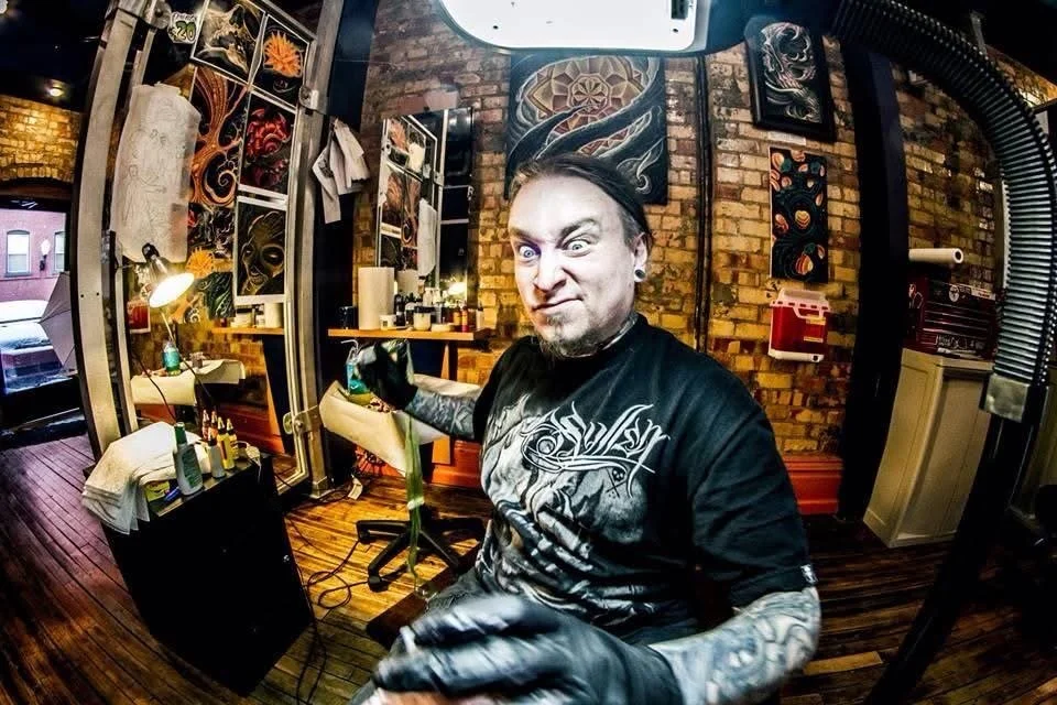 Man with tattoos and black gloves making a funny face in tattoo studio, with tattoo designs on brick wall behind him.