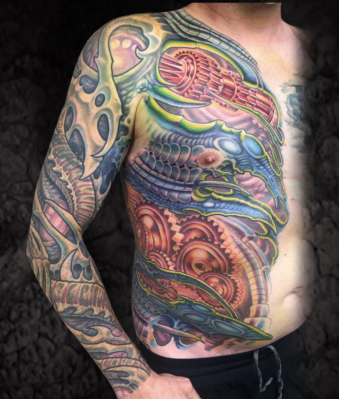 Colorful tattoo of a dragon covering the shoulder, chest, and arm of a man, featuring scales, gears, and mechanical elements.