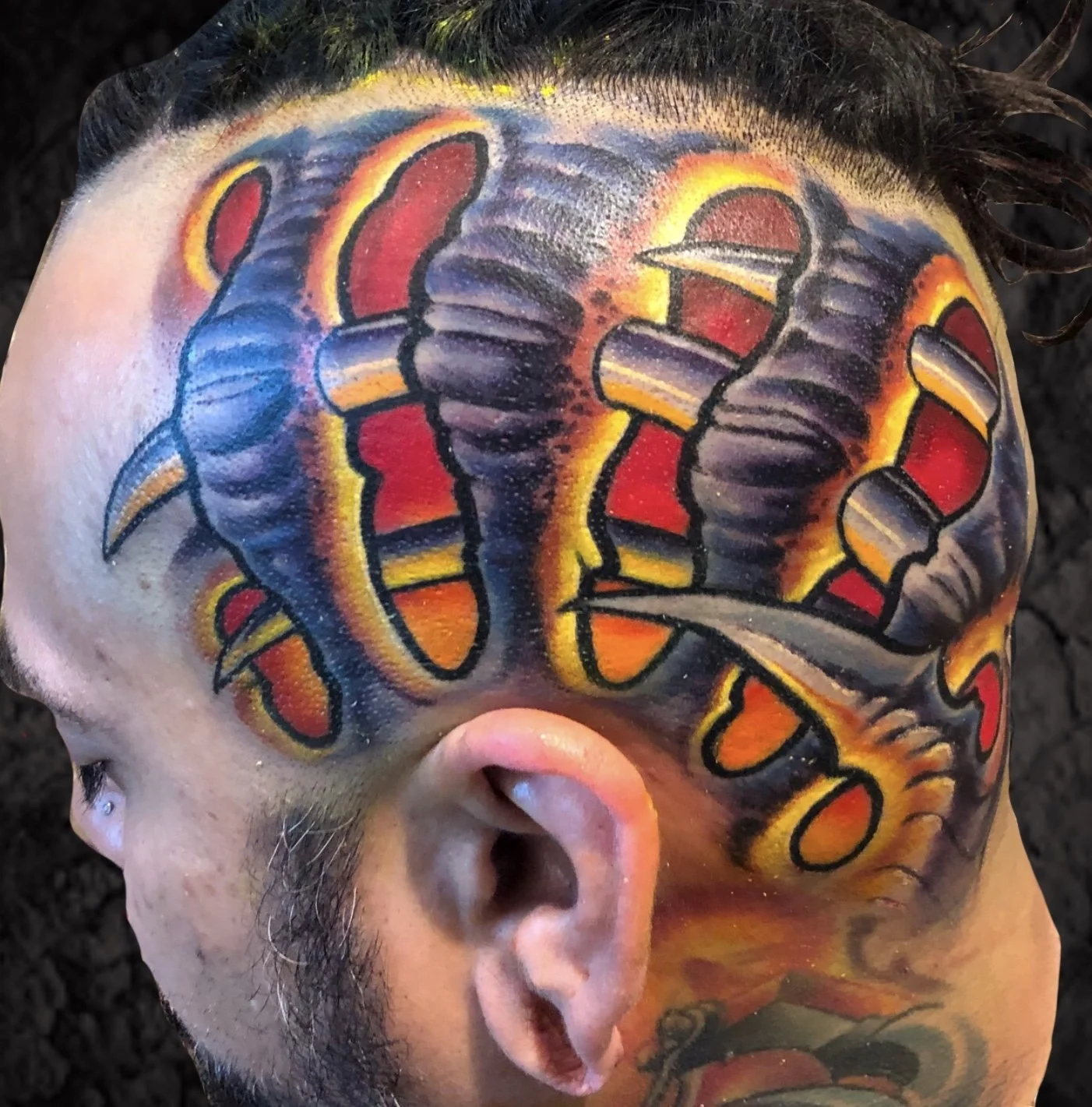 A person's head with a colorful tattoo of a snake wrapping around the skull, featuring red, yellow, orange, purple, and black ink.