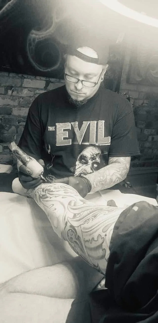 A man with glasses and tattoos on his arms tattooing a person's leg with elaborate designs.