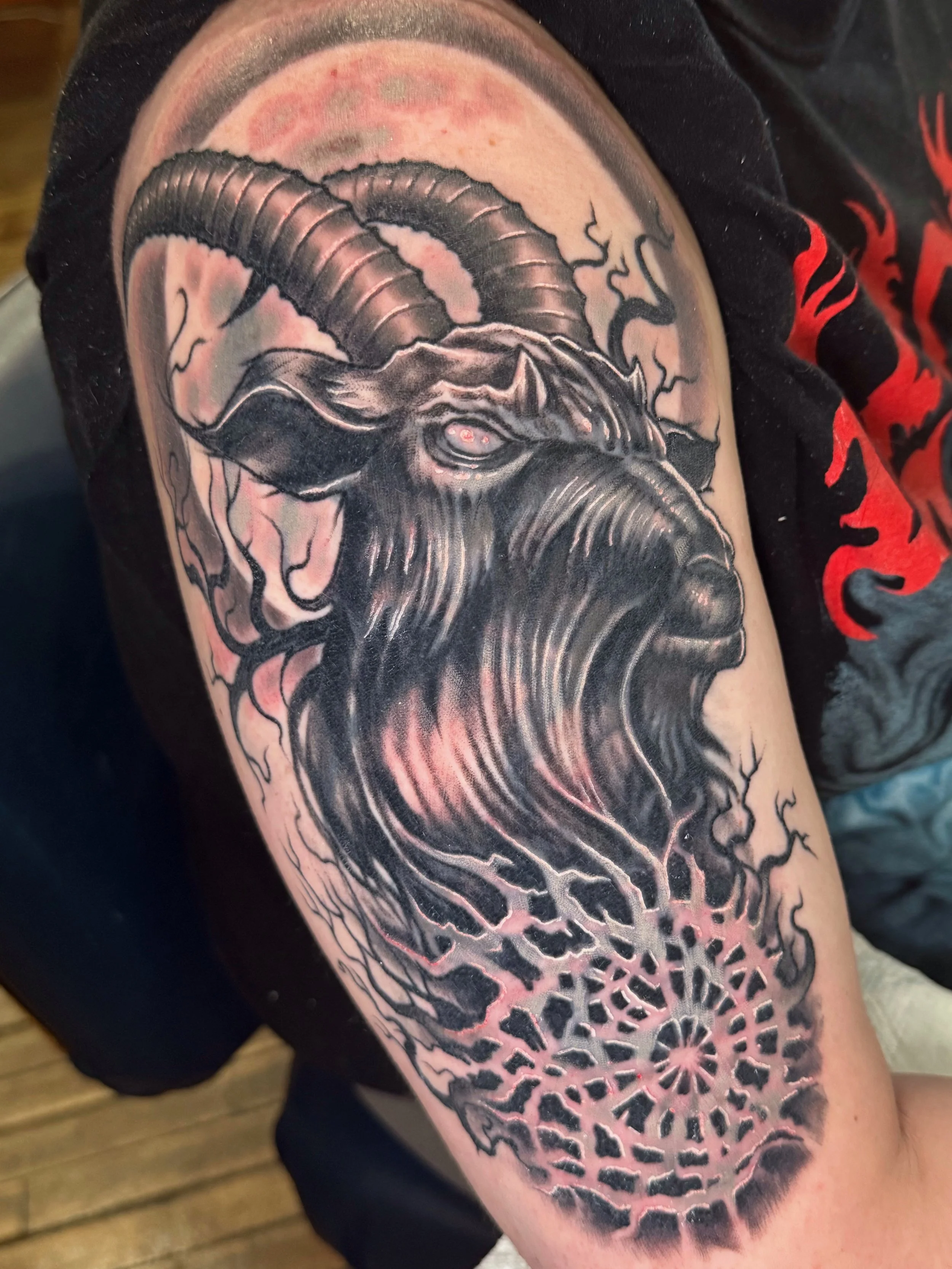 Tattoo of a goat with large horns and flowing beard, surrounded by a symmetrical geometric pattern, on a person's arm.