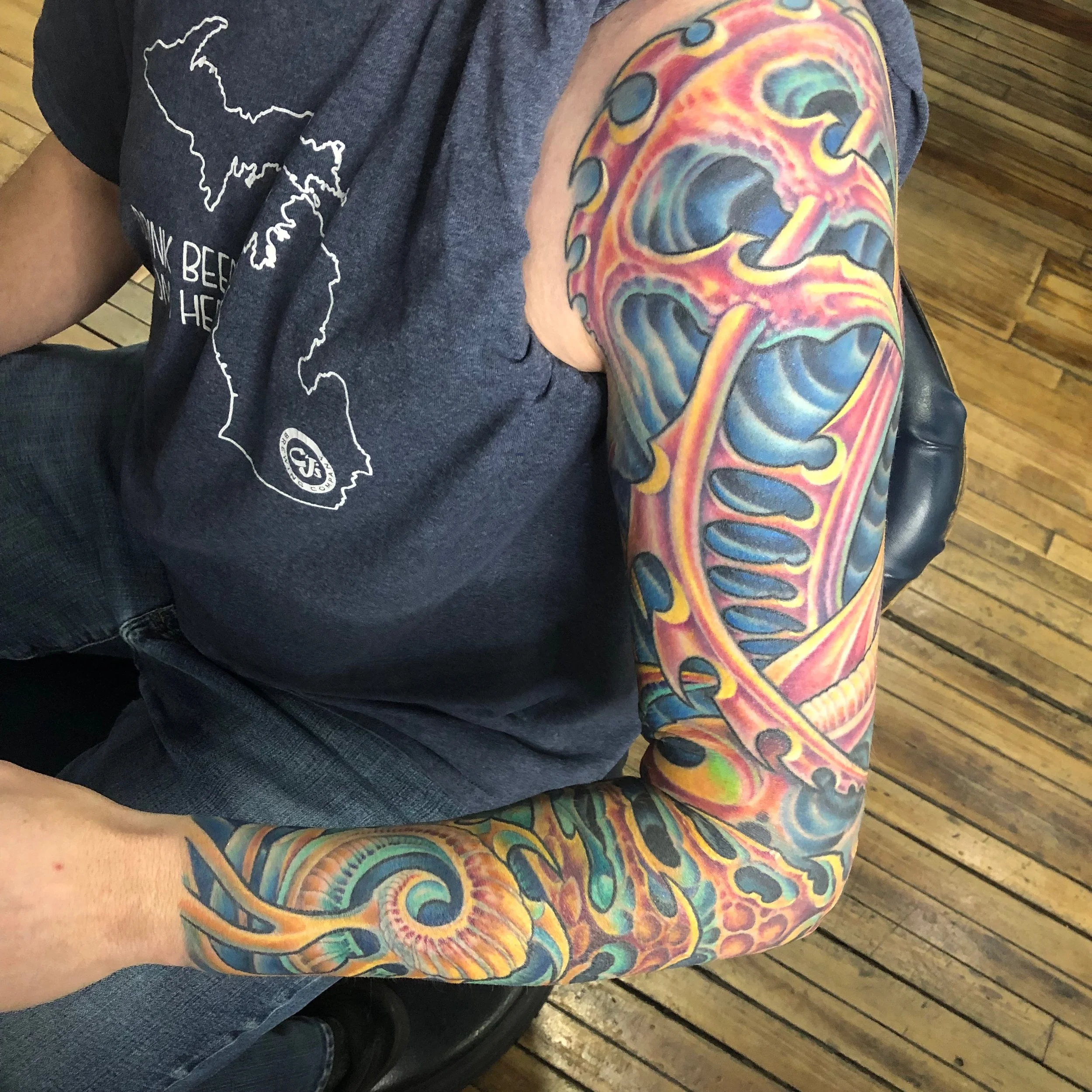 Close-up of a person's arm with a colorful, swirling tattoo sleeve featuring abstract patterns and bold hues of orange, blue, and purple, sitting on a wooden floor with a dark gray T-shirt nearby.