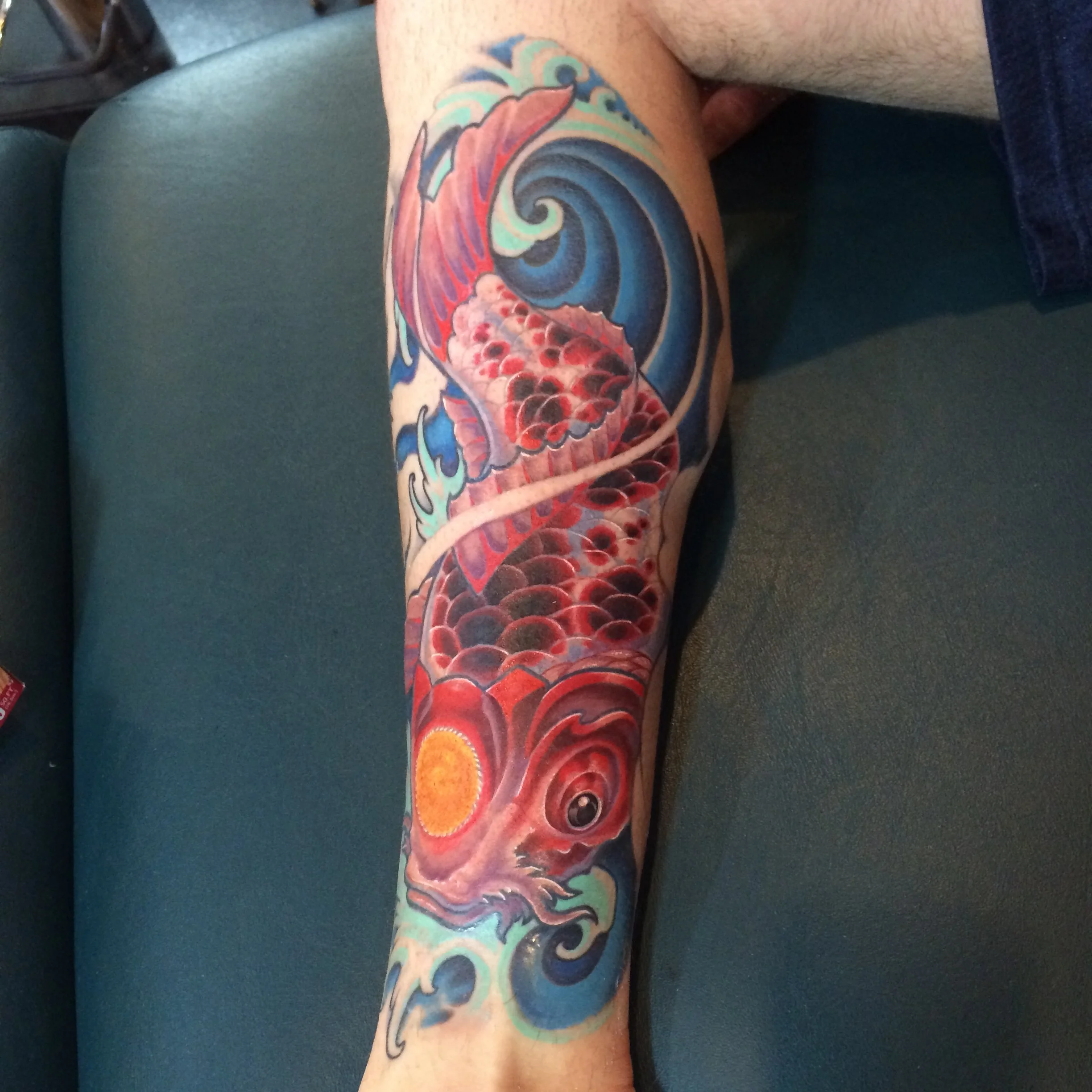 Colorful tattoo of a koi fish swimming in water, with blue and green waves and a bright yellow sun at the bottom of the arm.