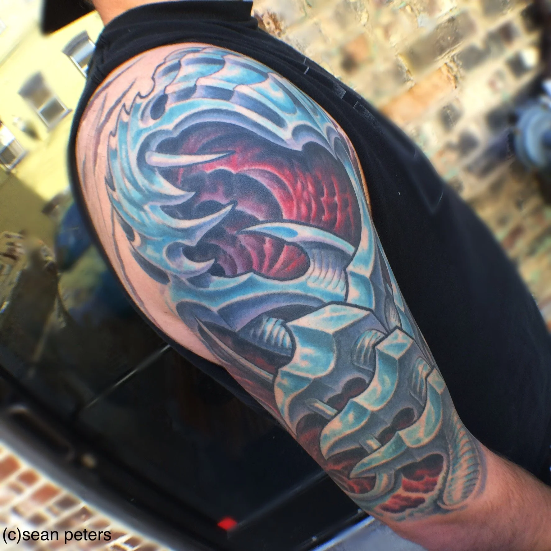 Colorful tattoo of a shark on a person's arm, black background, yellow building and brick pavement in the background.
