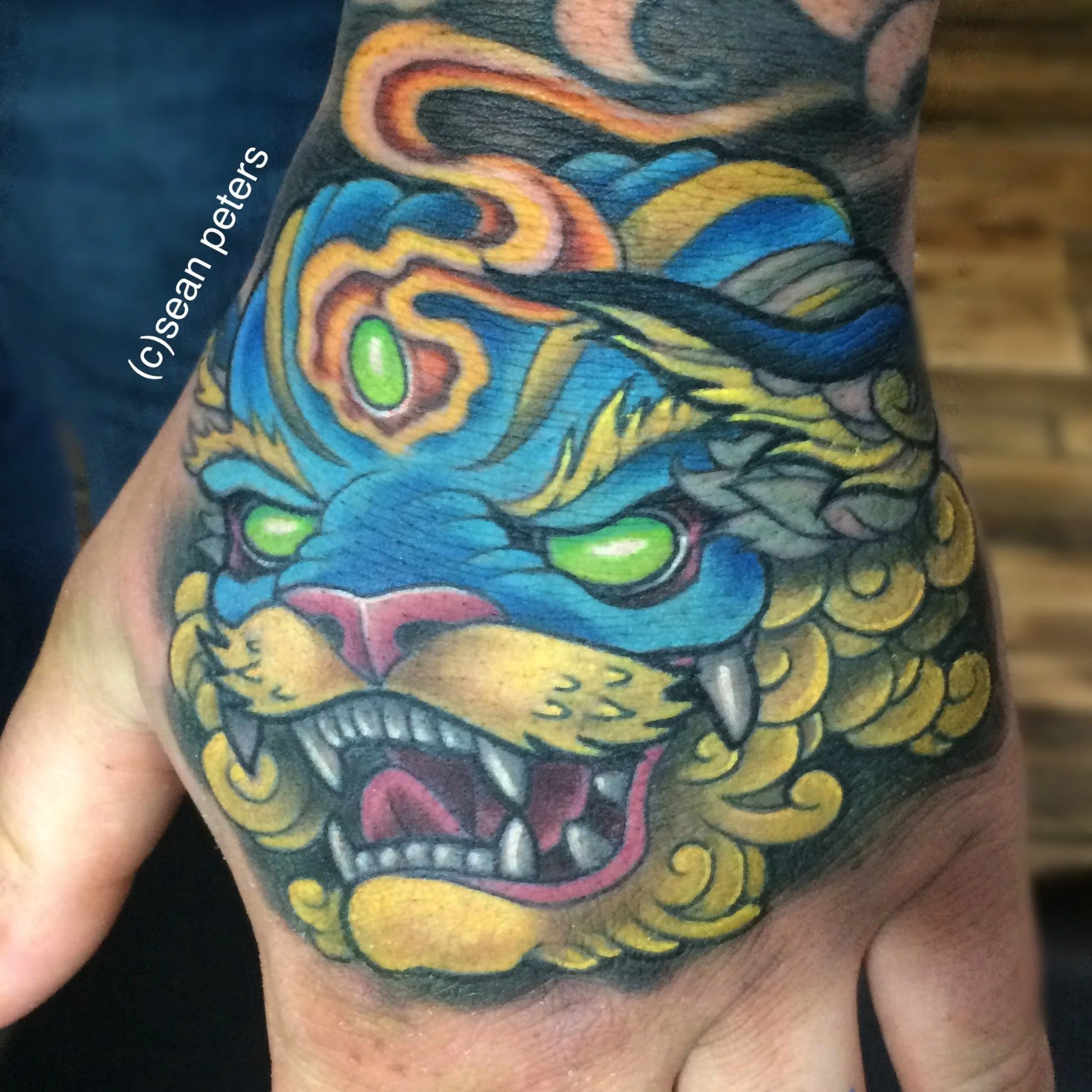 Close-up of a colorful tattoo of a dragon's face on a person's shoulder and upper arm. The dragon has vivid blue, yellow, orange, and green colors with detailed features.