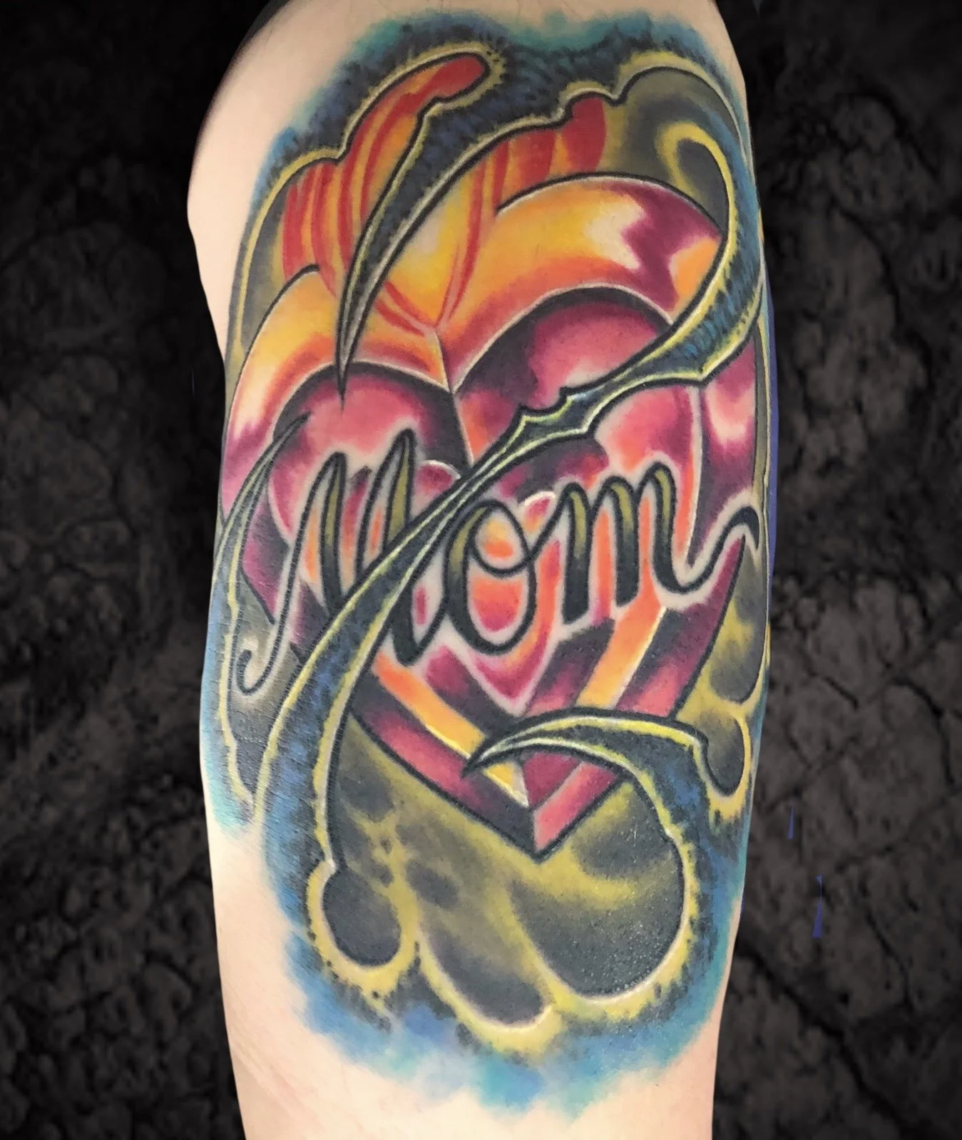 Colorful tattoo on a person's arm featuring the word "Mom" surrounded by floral and abstract designs in red, yellow, green, blue, and black ink.