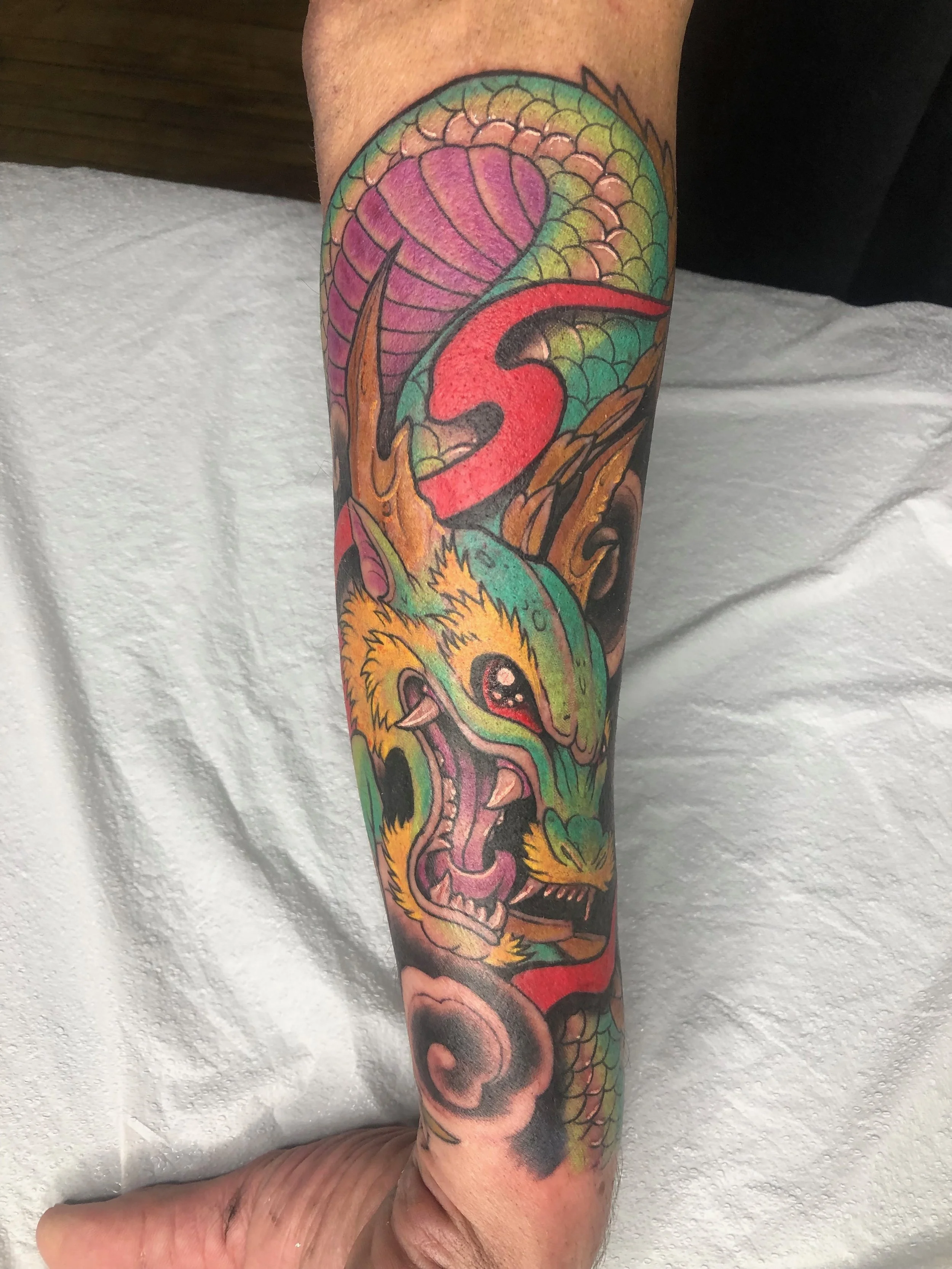 Colorful tattoo of a dragon's head on a person's arm, featuring scales, flames, and vibrant colors like pink, green, and yellow.