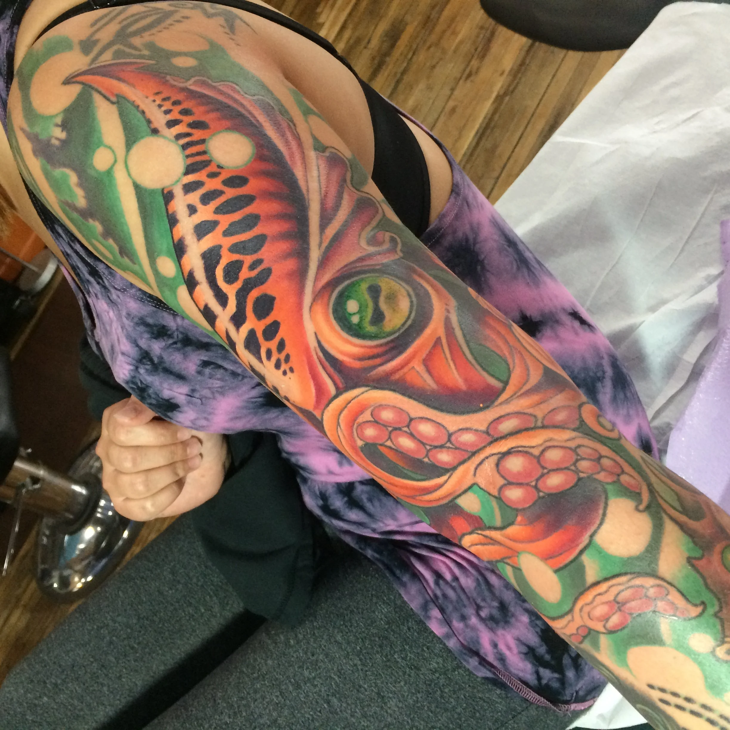 Colorful tiger and animal face tattoo covering the person's arm and shoulder, with the person sitting next to a table and holding their hand.