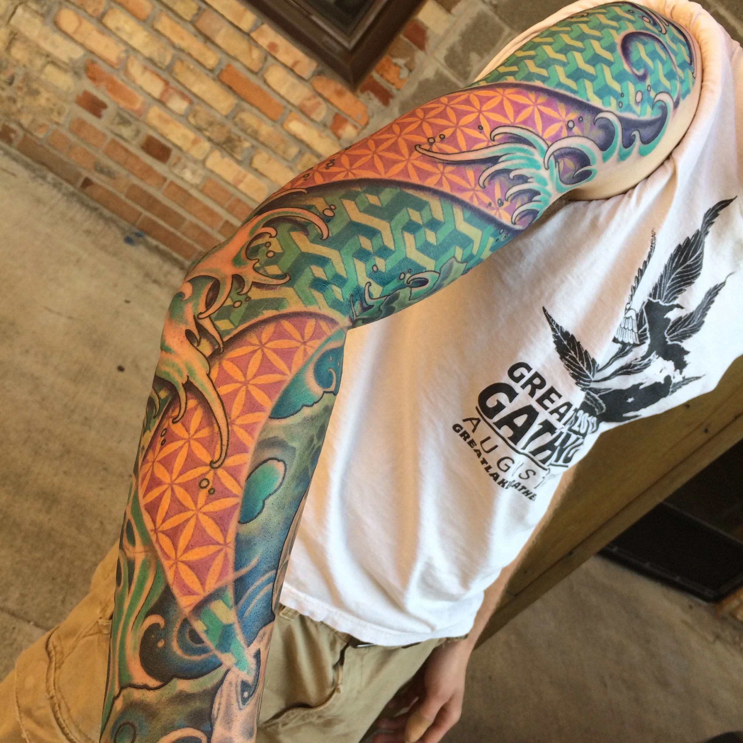 A person with a colorful tattoo sleeve featuring geometric patterns, waves, and abstract designs on their arm, wearing a white t-shirt with black print, standing near a brick fireplace.