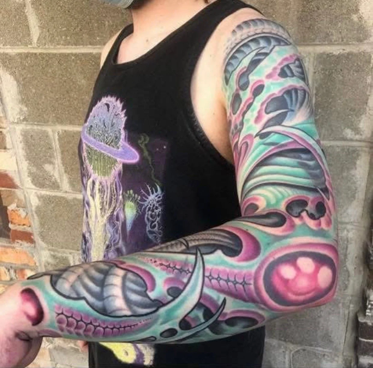 A person with colorful tattoos on their arm, wearing a black tank top with a space-themed design, standing against a brick and concrete wall.