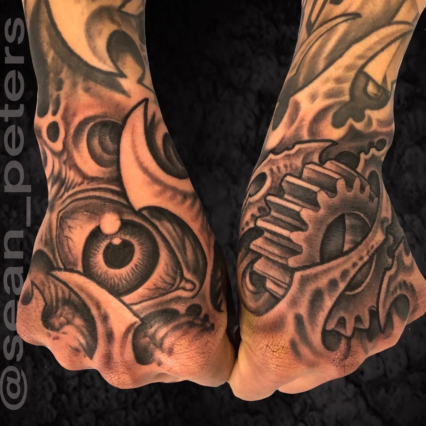 Close-up of tattooed forearms with black and gray detailed designs, including an eye, gears, and abstract patterns.