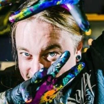 Person with light-colored hair and blue eyes wearing black gloves and holding a tie-dye colored object above their head.