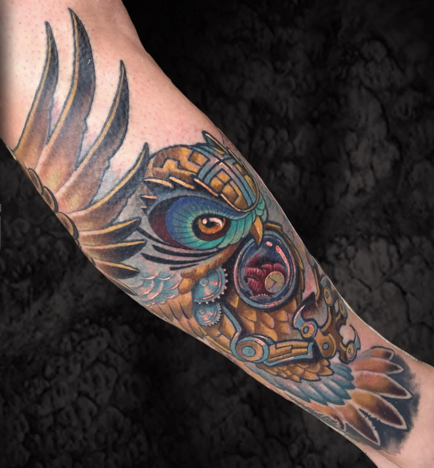 Colorful tattoo of a bird with intricate design on a person's arm, showing black outlining and vibrant shades of blue, green, orange, and pink, against a dark background.