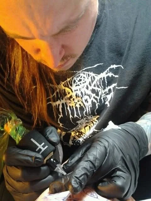 A person with reddish hair and a gray T-shirt, wearing black gloves, is using a tattoo machine to ink a design on someone's skin.