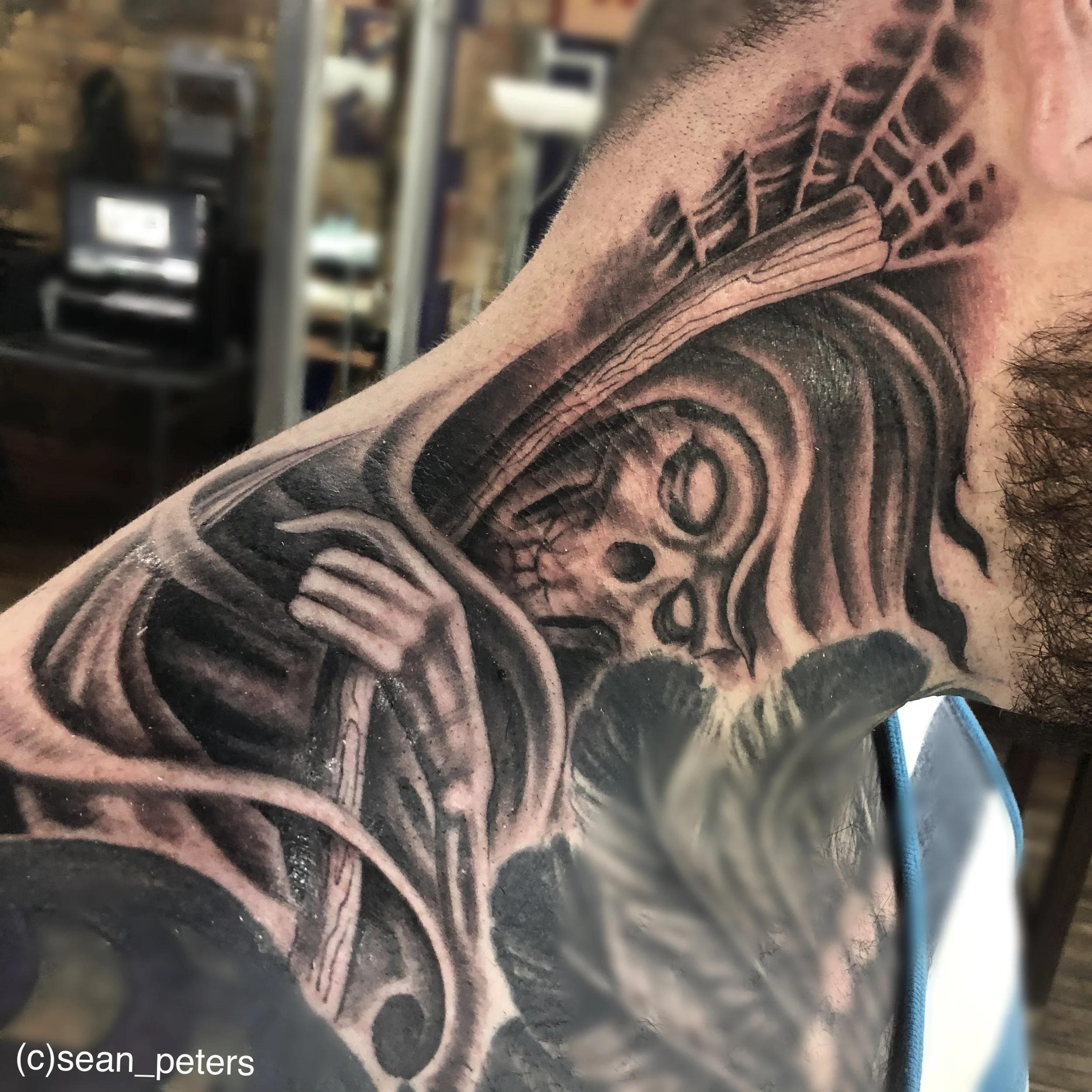 A close-up image of a detailed tribal-style tattoo on a person's upper arm and shoulder, partially covered with hair. The tattoo features bold black lines and intricate patterns.