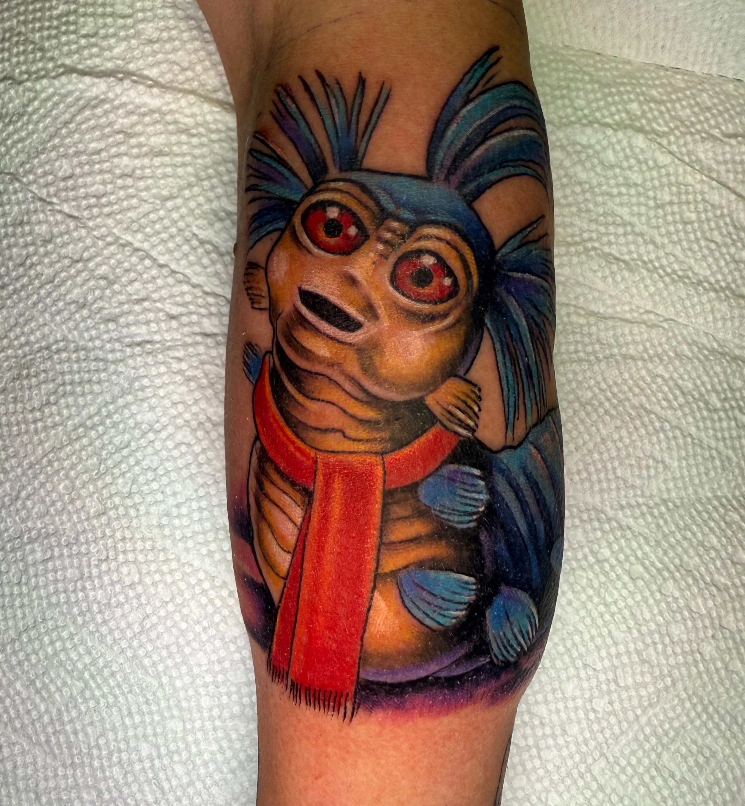 Colorful tattoo of a clown with red eyes, yellow face, and blue hair on a person's arm, with a red nose and a black smile, on a white textured background.