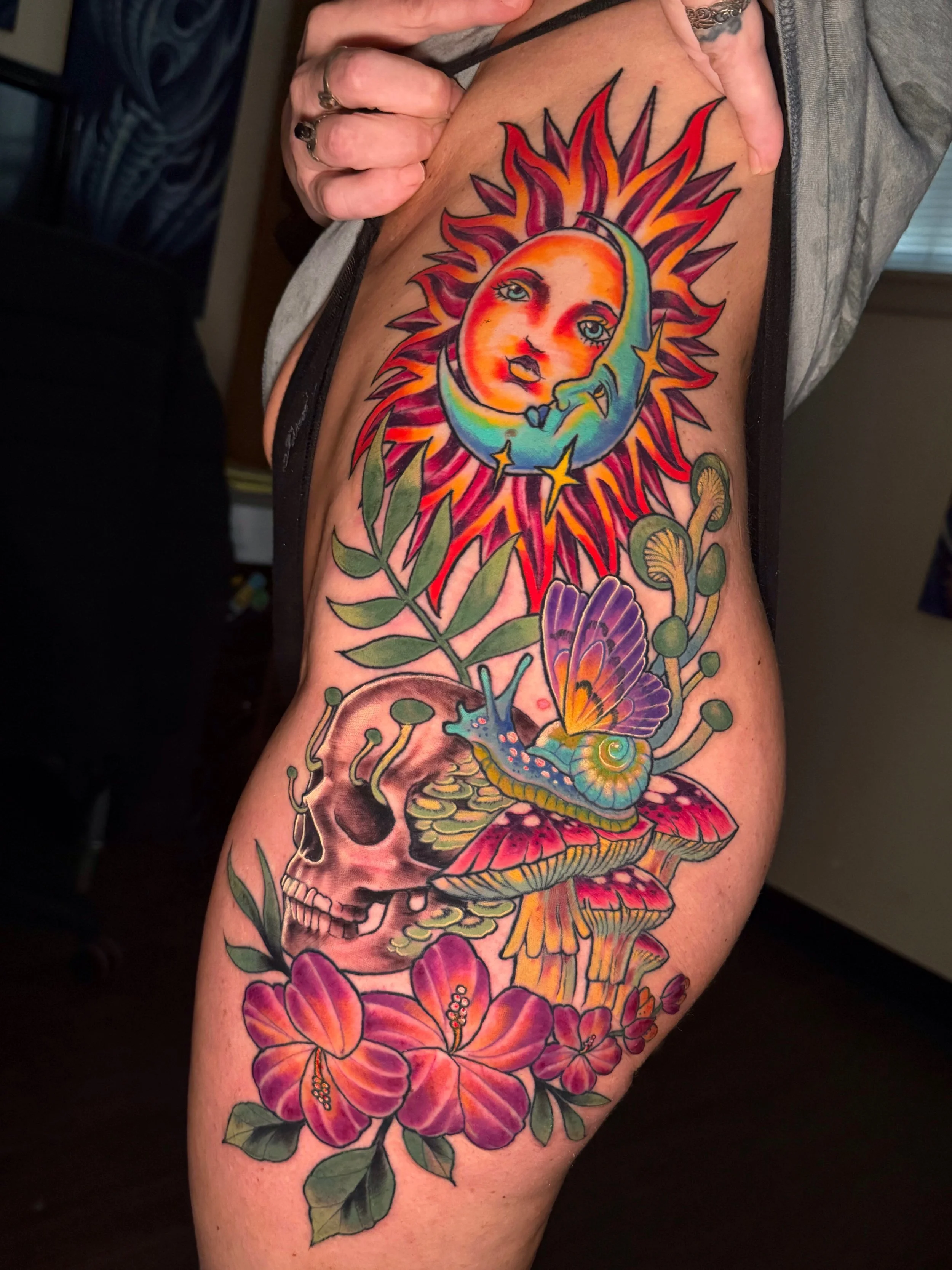 Colorful tattoo of a sun with a woman's face in the center, surrounded by flowers, leaves, a skull, and a snail, on a person's arm.