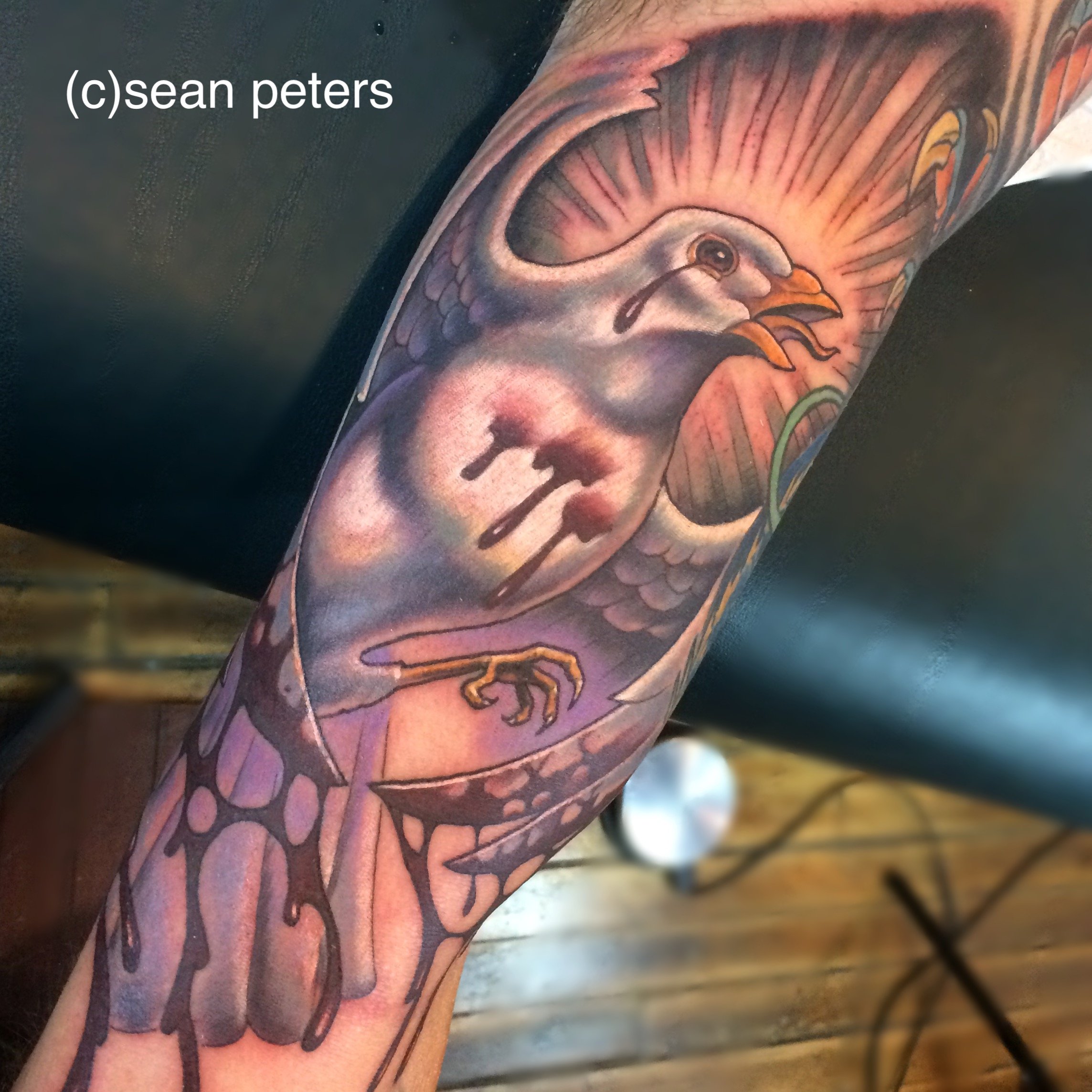 Close-up of a tattooed arm featuring a traditional style bird with detailed feathers and a beak, surrounded by colorful elements and black outlines.