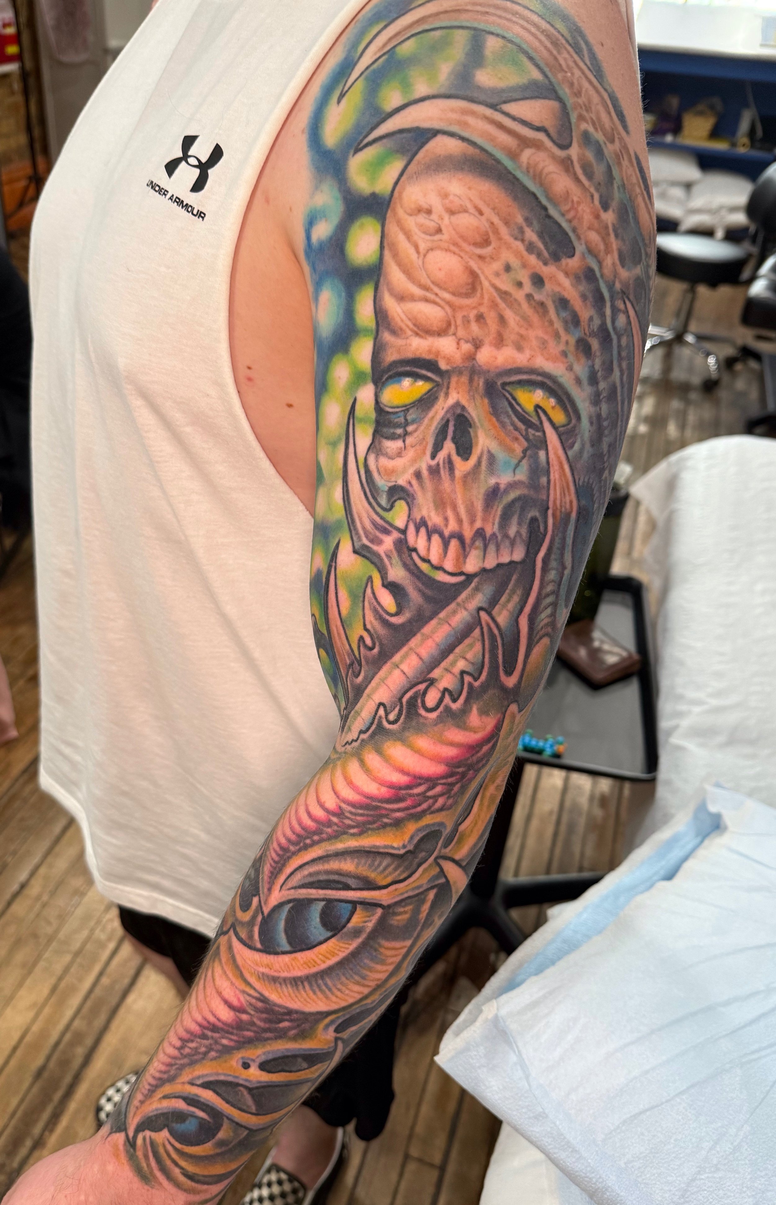 A colorful tattoo of a sinister clown skull with glowing yellow eyes, detailed with vivid greens, blues, and reds, covering the person's upper arm.