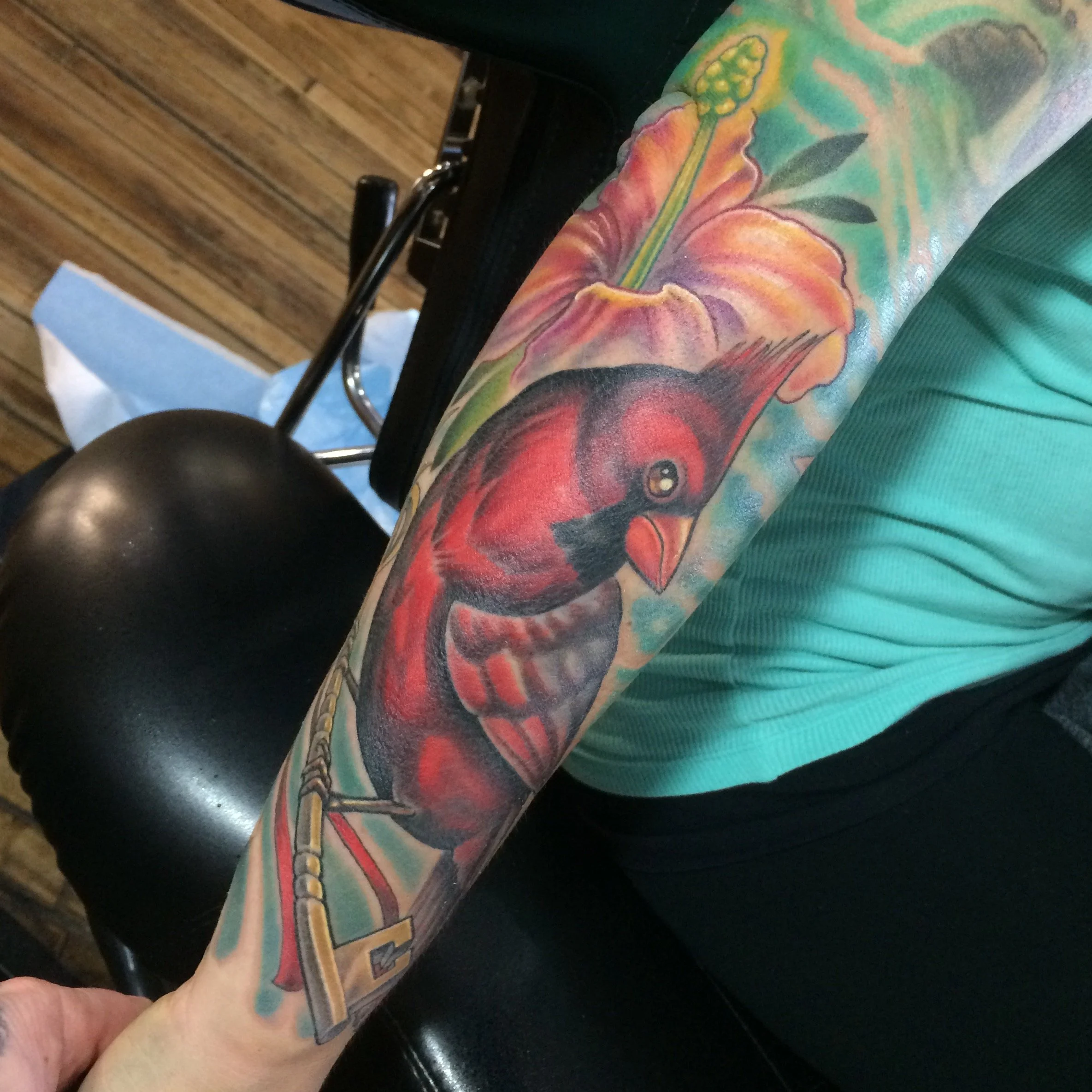 Close-up of a person's arm with a colorful tattoo of a hummingbird and floral design, wearing a teal sleeve.