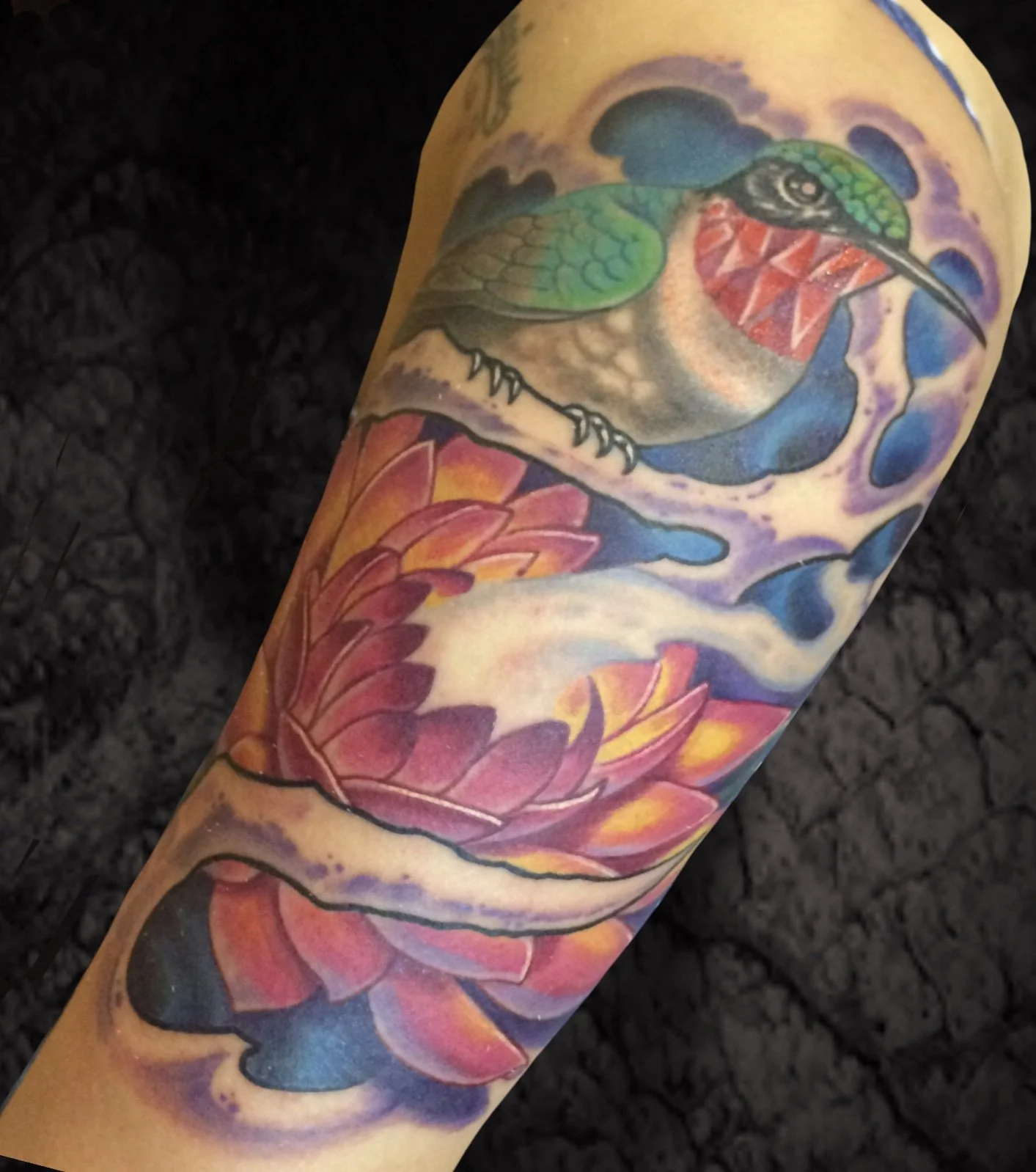 Colorful tattoo of a hummingbird with green feathers and red chest, perched on a branch adorned with pink and orange flowers, set against a background of purple and blue clouds.