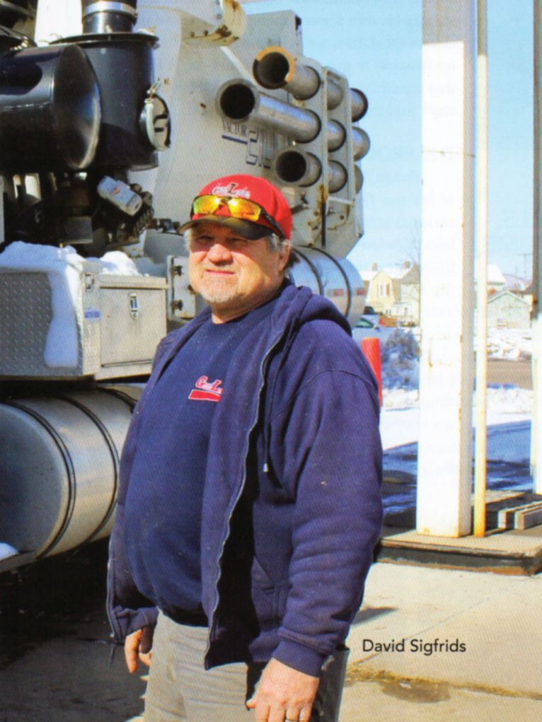 A man wearing a red cap, sunglasses, a navy blue hoodie, and a navy blue sweatshirt standing next to a large industrial machine outdoors on a sunny day.