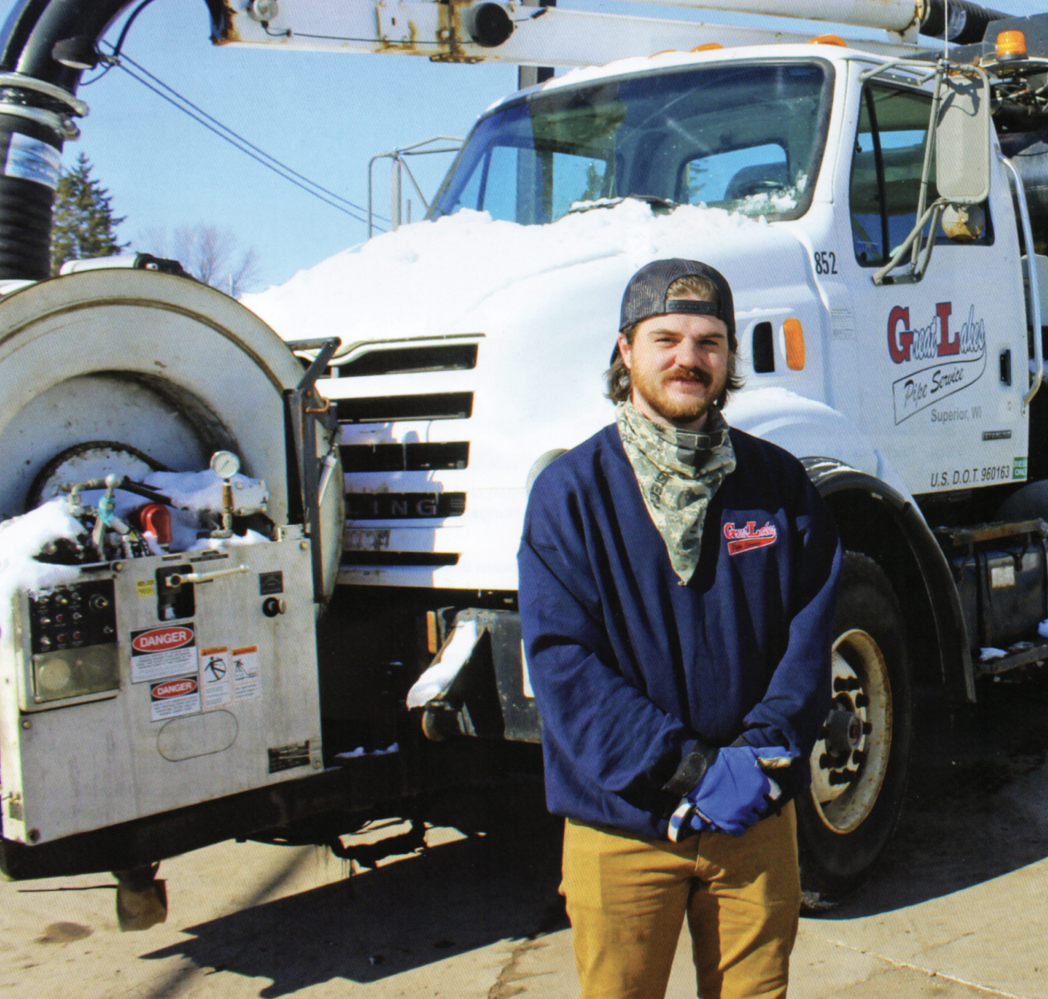 A man with a beard, wearing a backward baseball cap, a blue jacket with a logo, tan pants, and blue gloves, standing in front of a utility truck with snow on it.