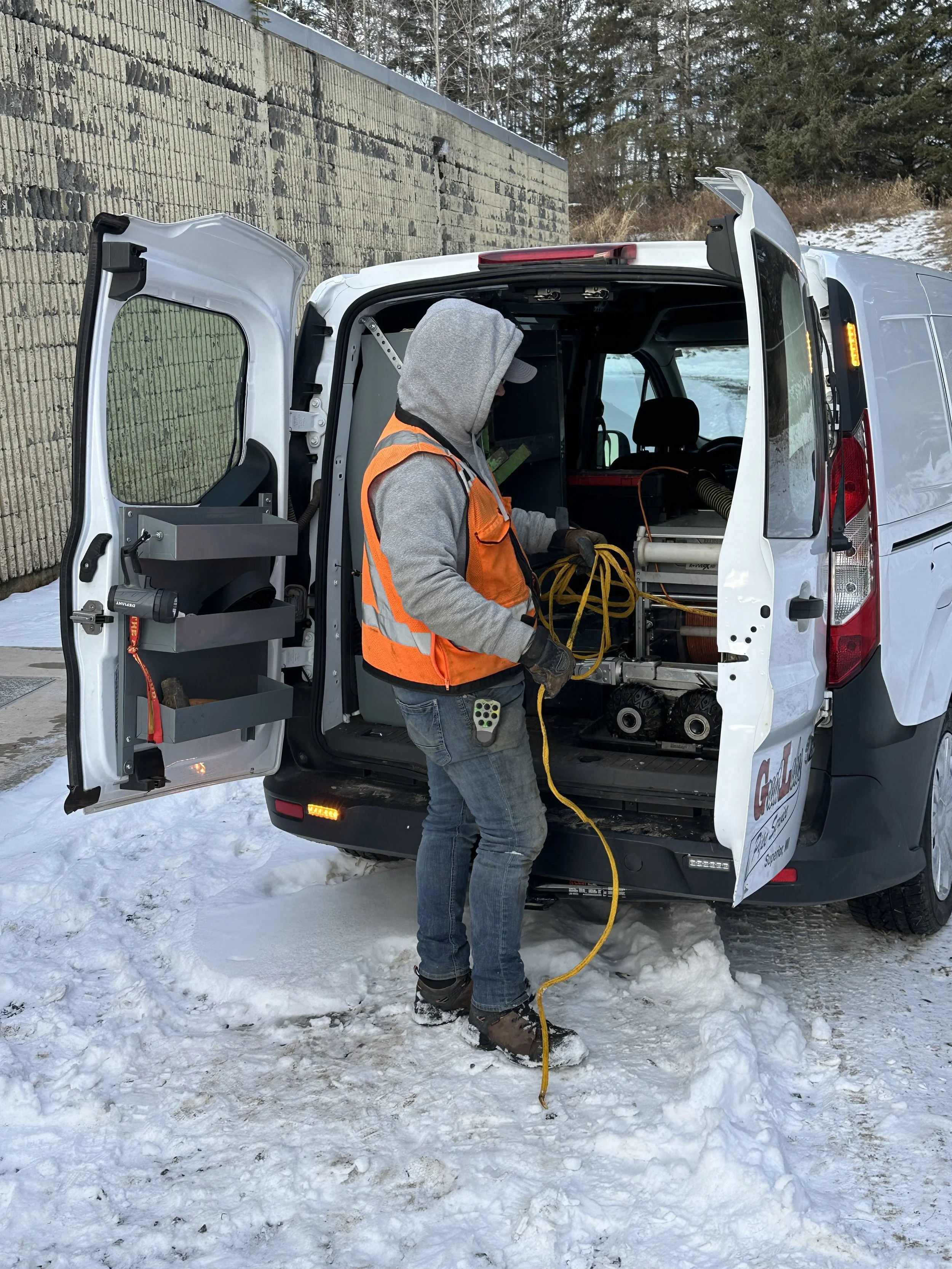 A person wearing a gray hoodie, orange safety vest, jeans, and winter boots working with equipment in the back of a white service van on snow-covered ground. The person is holding a yellow cord and appears to be installing or repairing something inside the van, which contains various tools and machinery. The background shows a stone wall and trees with some snow.