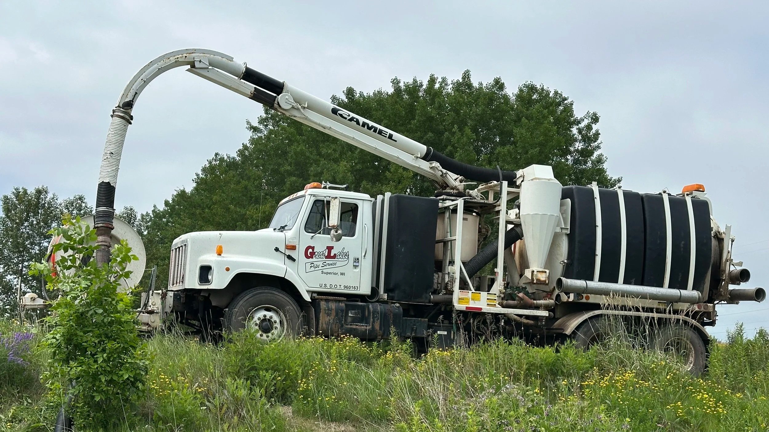 A large white vacuum truck with black hoses and orange warning lights is parked on grassy land with trees in the background.