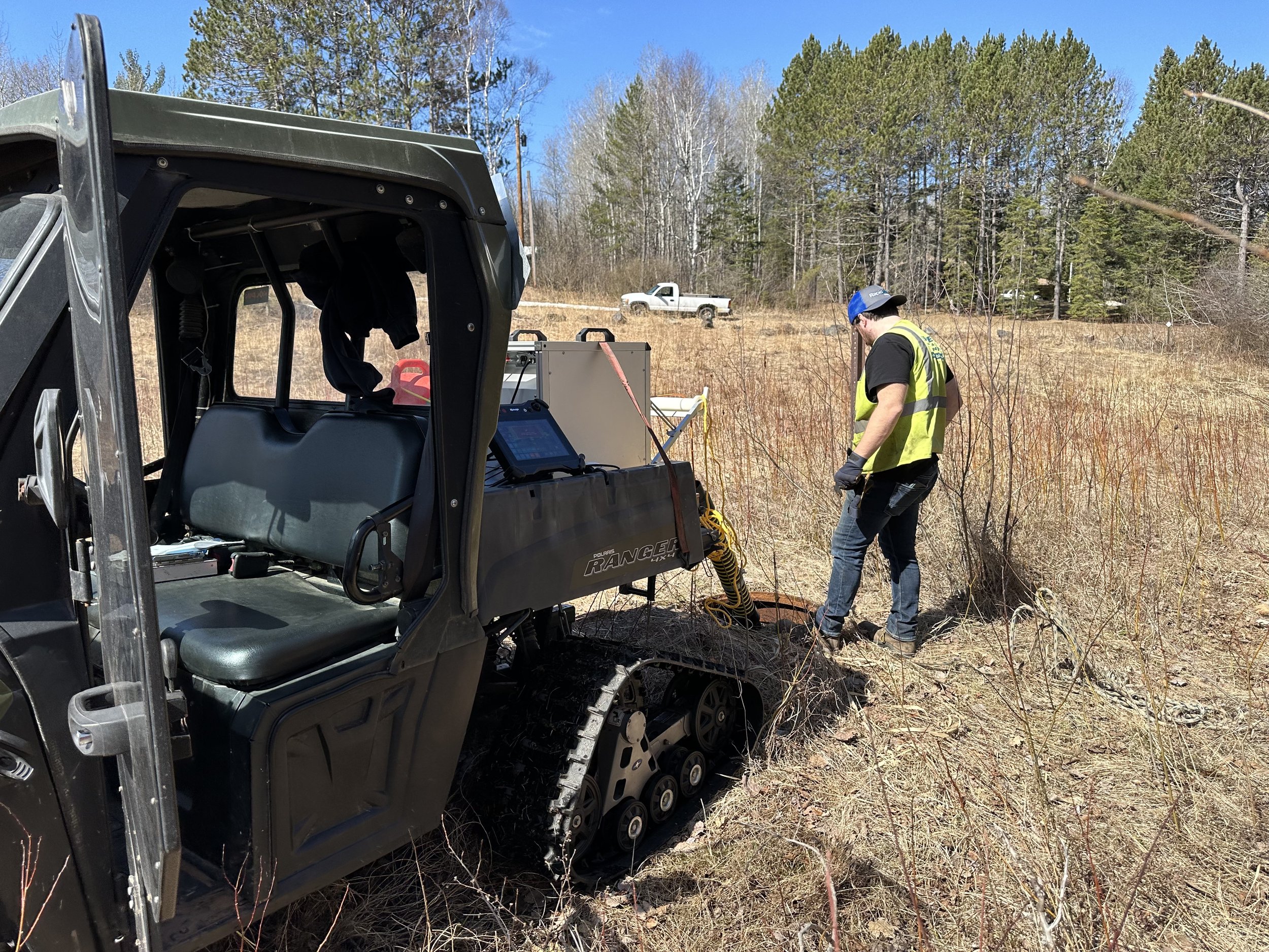 A person in a safety vest and cap working outdoors in a grassy, wooded field next to a tracked utility vehicle. There is equipment on a table and a white pickup truck in the background under a blue sky.