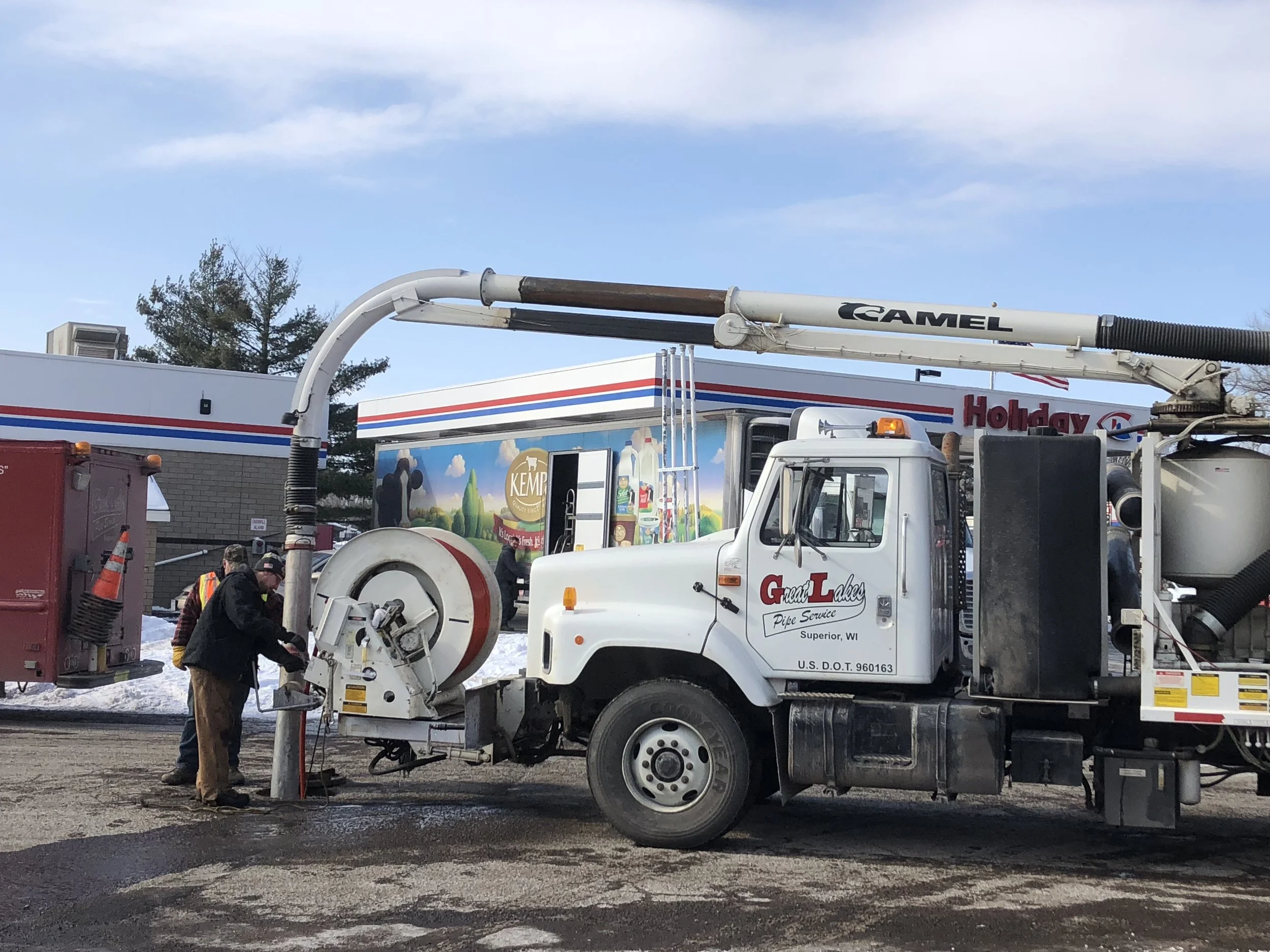 Workers repairing a large pipe connected to a utility truck at a service site, with a Holiday gas station sign and a blue sky with clouds in the background.