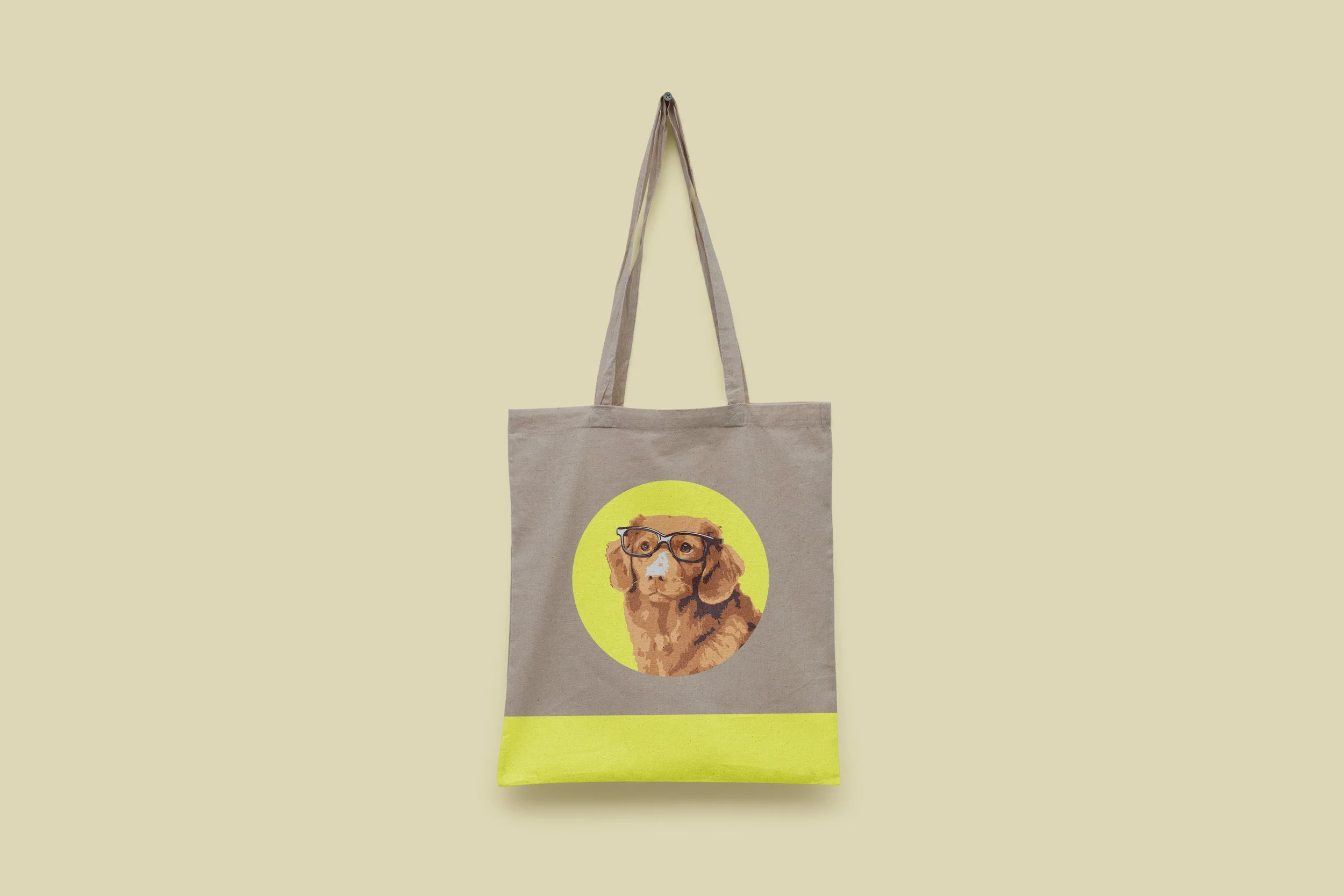A beige tote bag with a graphic of a dog wearing glasses printed on it, hanging against a plain background.