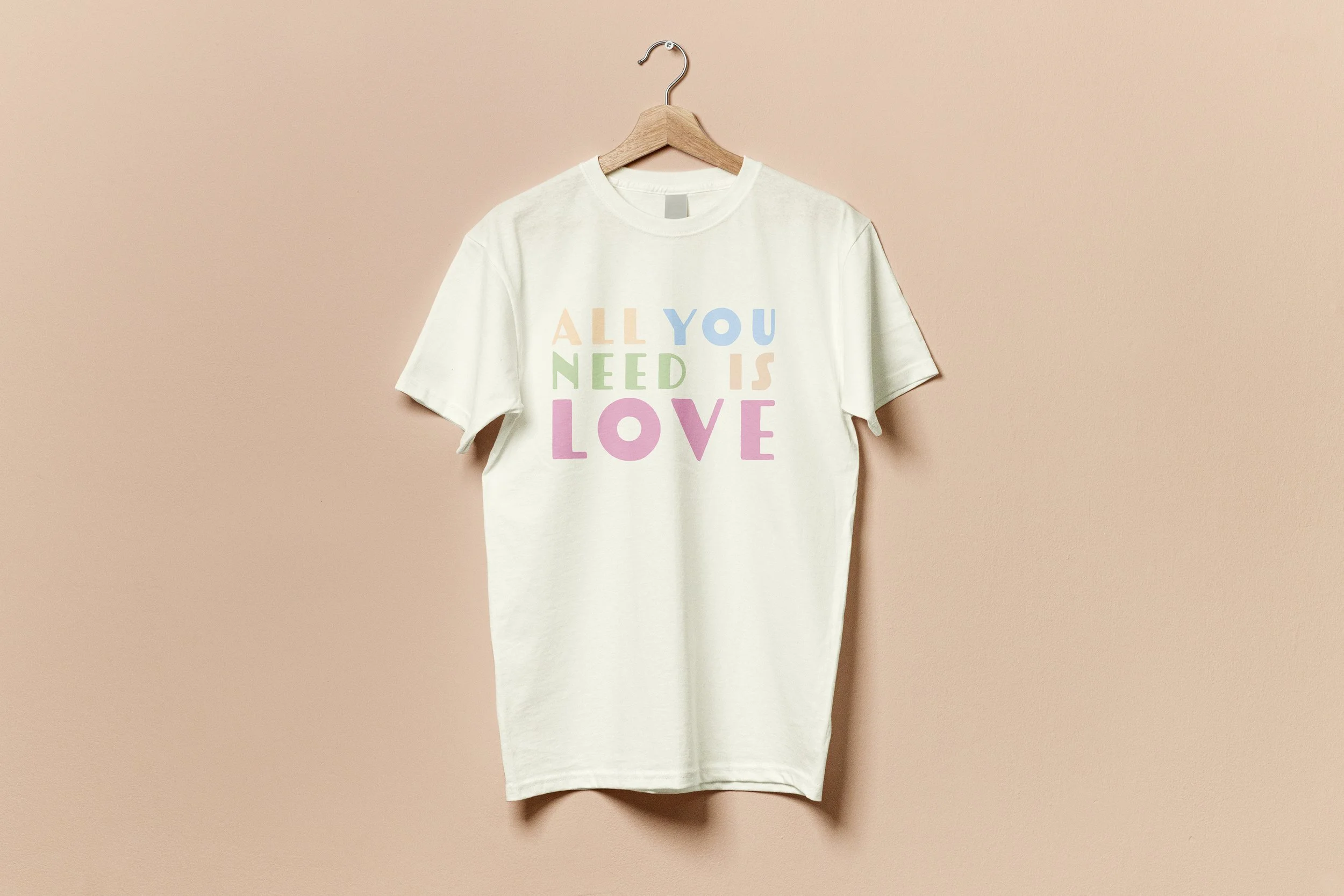 A white T-shirt with colorful pastel lettering that reads 'ALL YOU NEED IS LOVE,' hanging on a wooden hanger against a pink wall.
