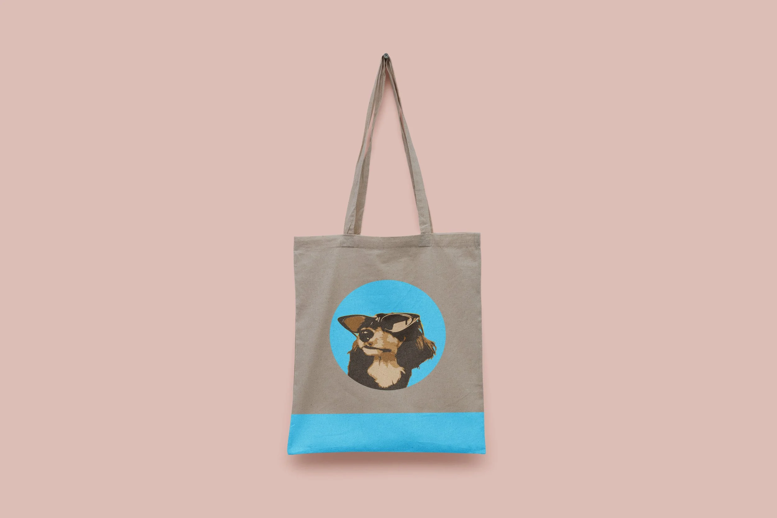 A beige tote bag with a circular graphic of a dog wearing glasses and a blue background, hanging against a pink background.