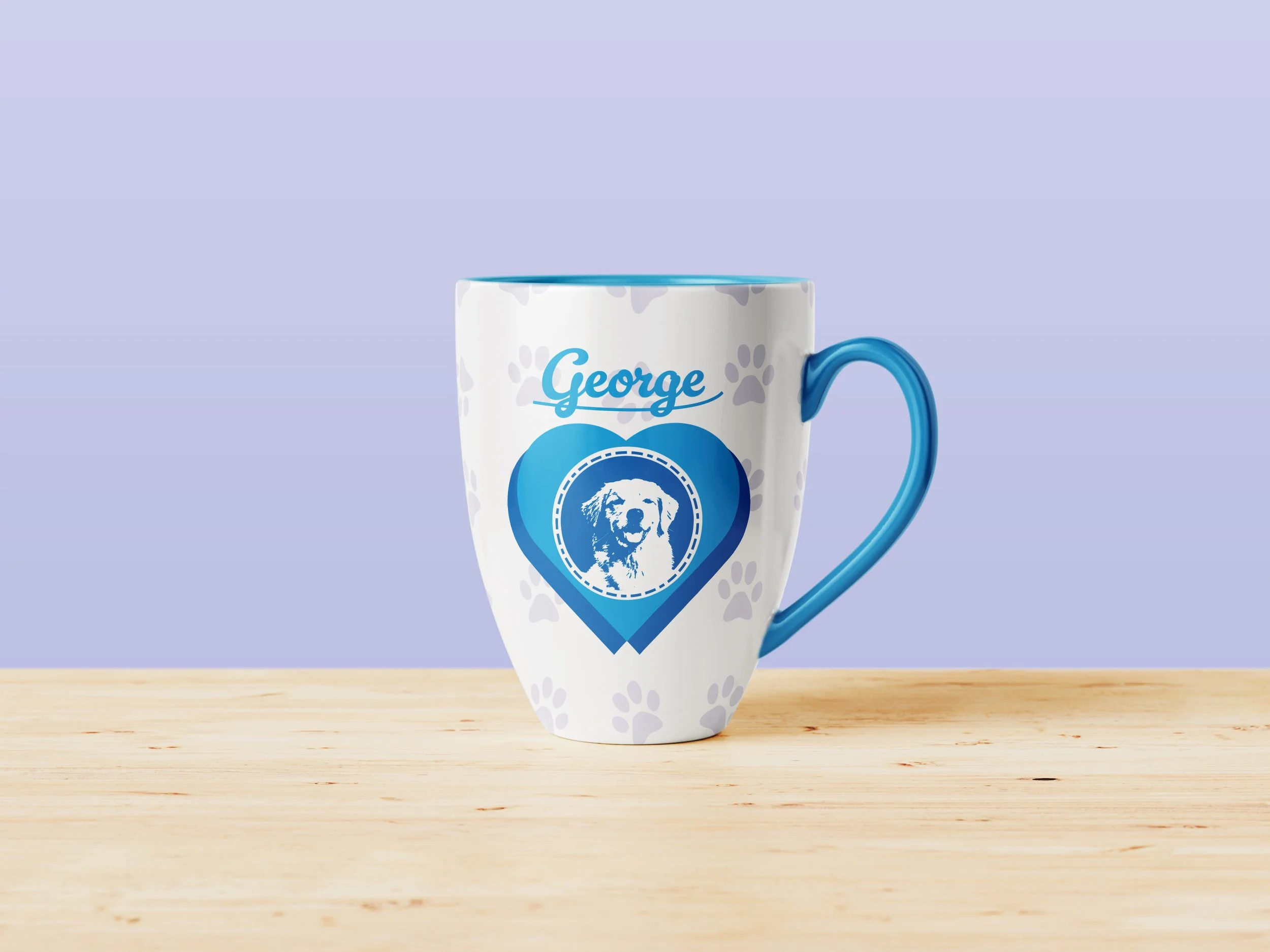 White coffee mug with light gray paw print pattern, blue handle, and blue inner rim, featuring a personalized name "George" and a dog graphic on the front, placed on a wooden surface with a pastel purple background.