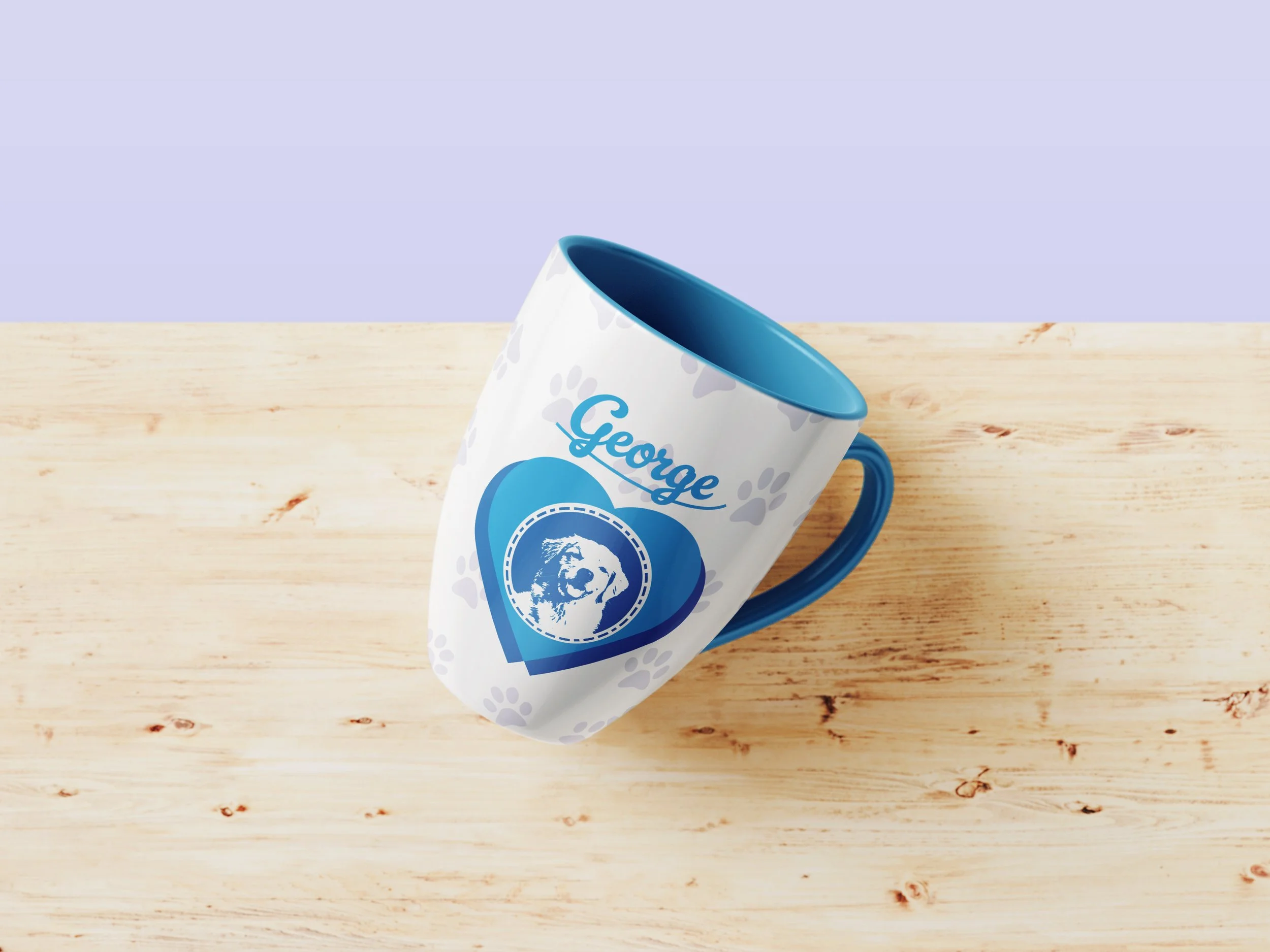 A white ceramic mug with a blue handle and interior, sitting on a light wooden surface. The mug has a design featuring a blue heart, a dog silhouette, and the word 'George' written above the heart.