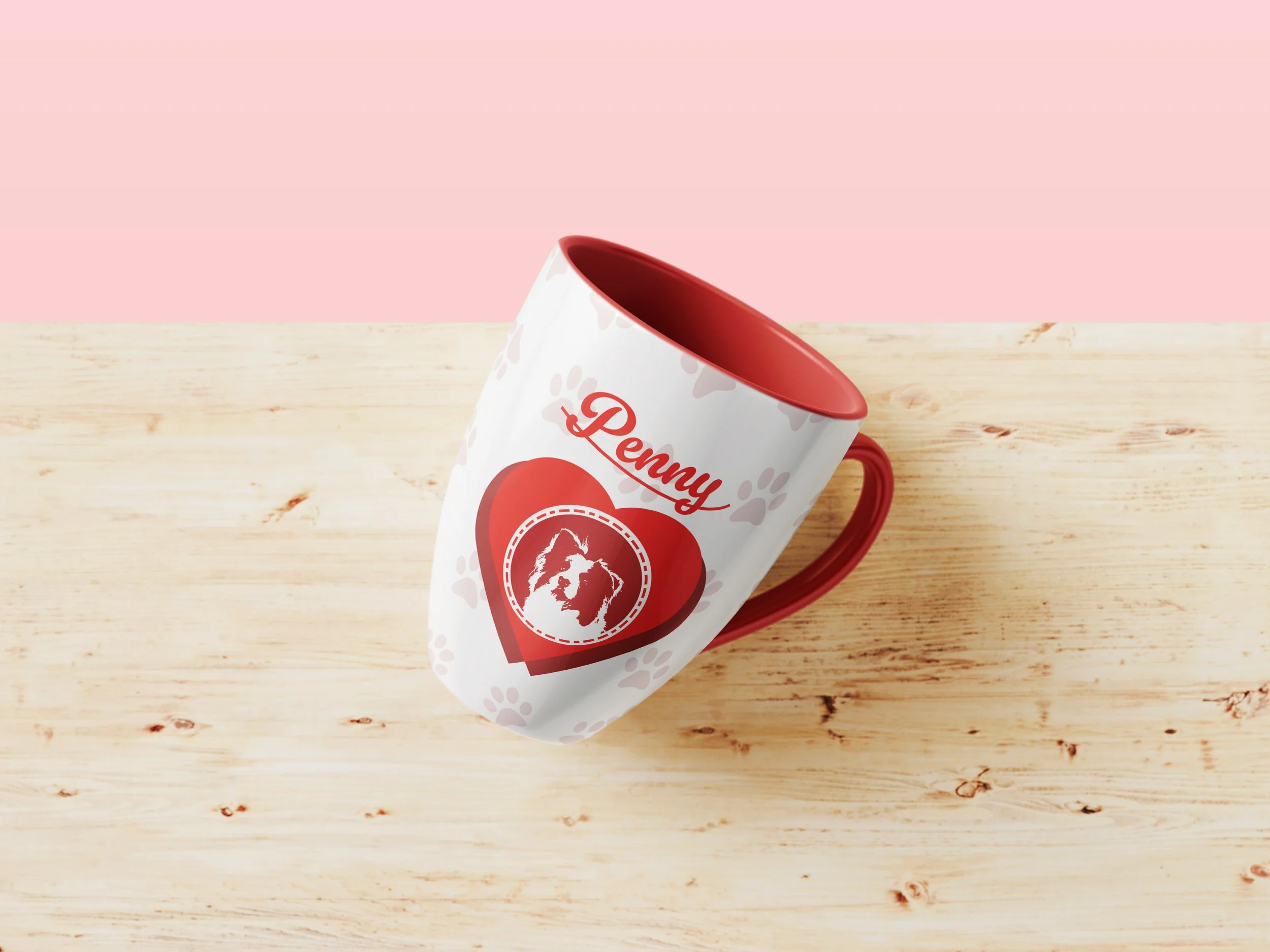 A white coffee mug with a red interior and red handle, featuring a dog and the word 'Penny' printed on it, placed on a light wooden surface against a light pink background.