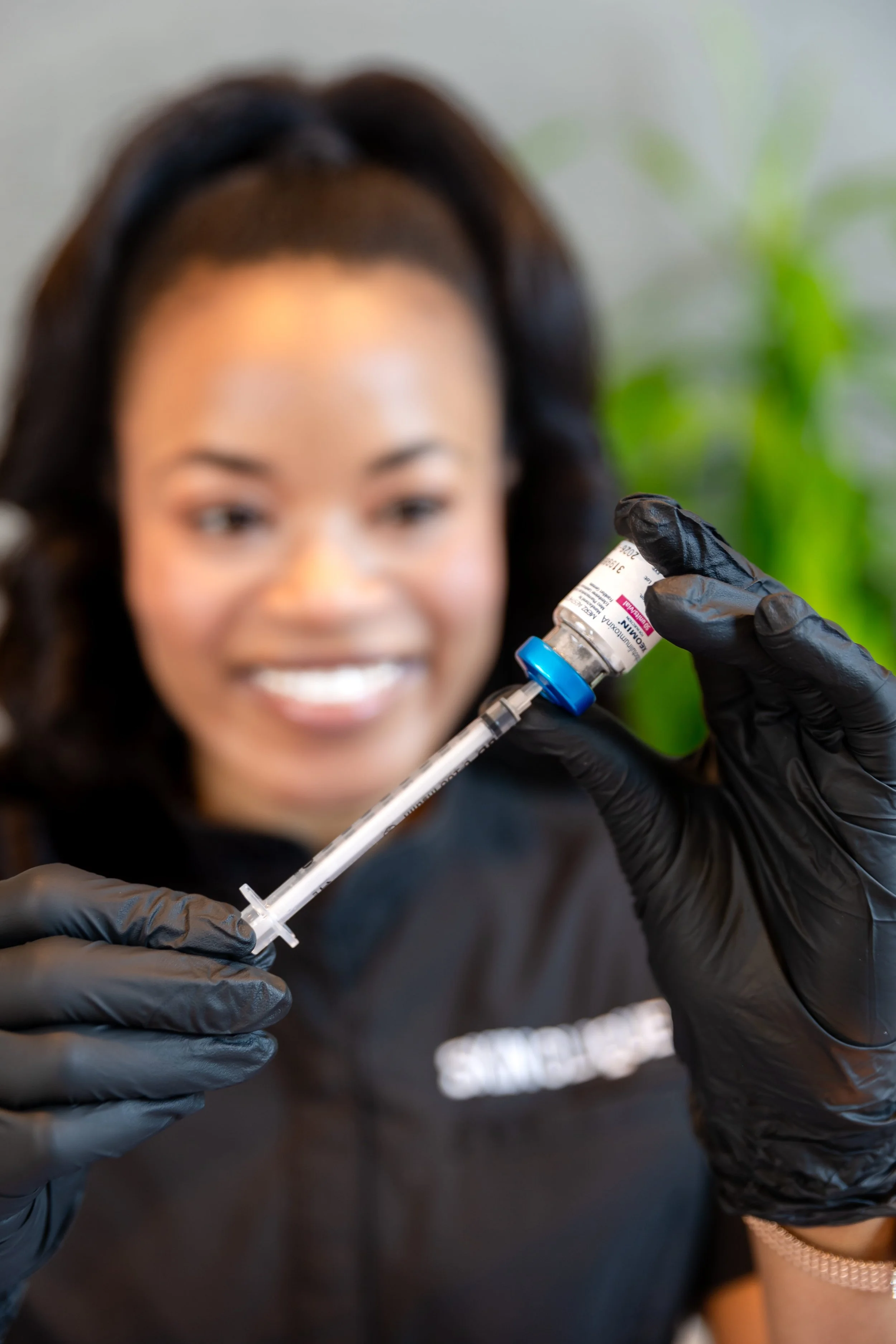 A woman with dark hair and a smile preparing to receive a vaccination, wearing black gloves, with a vaccine vial and syringe in focus.