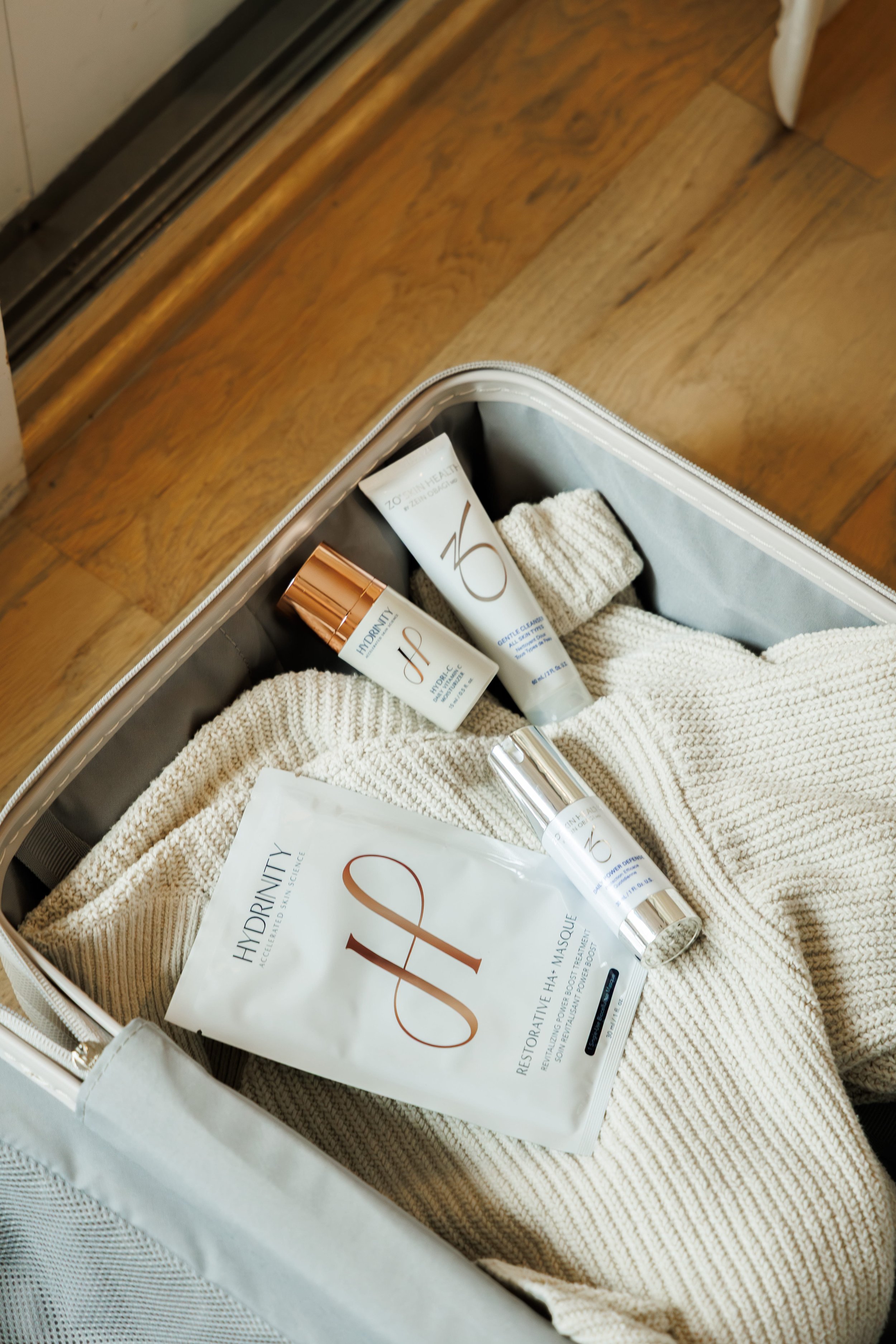 Open bag containing skincare products, a small cream-colored knit towel, and a white packet labeled 'Hydrinity' on a wooden floor.