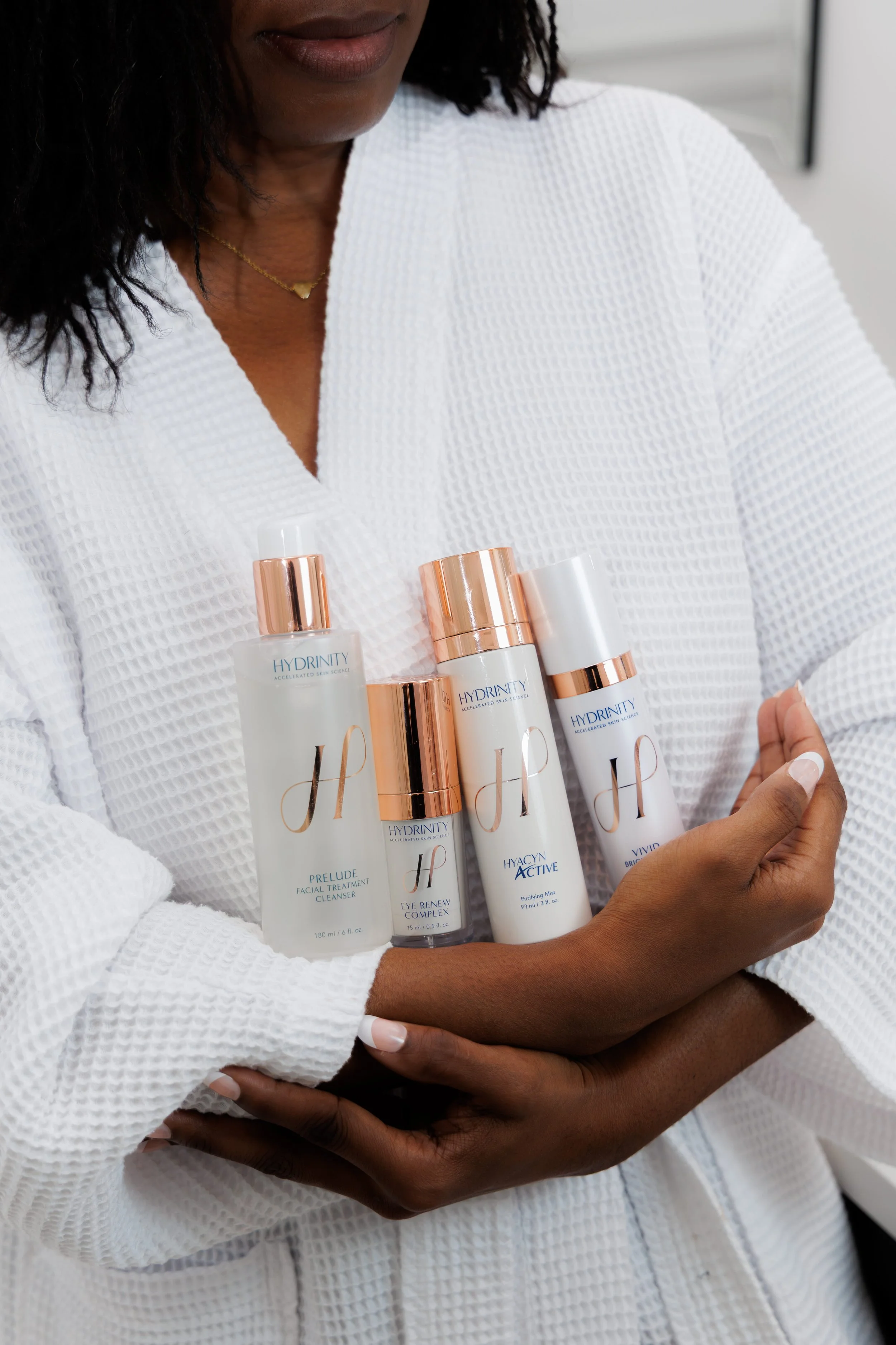 A woman in a white textured robe holding five skincare bottles with gold caps.