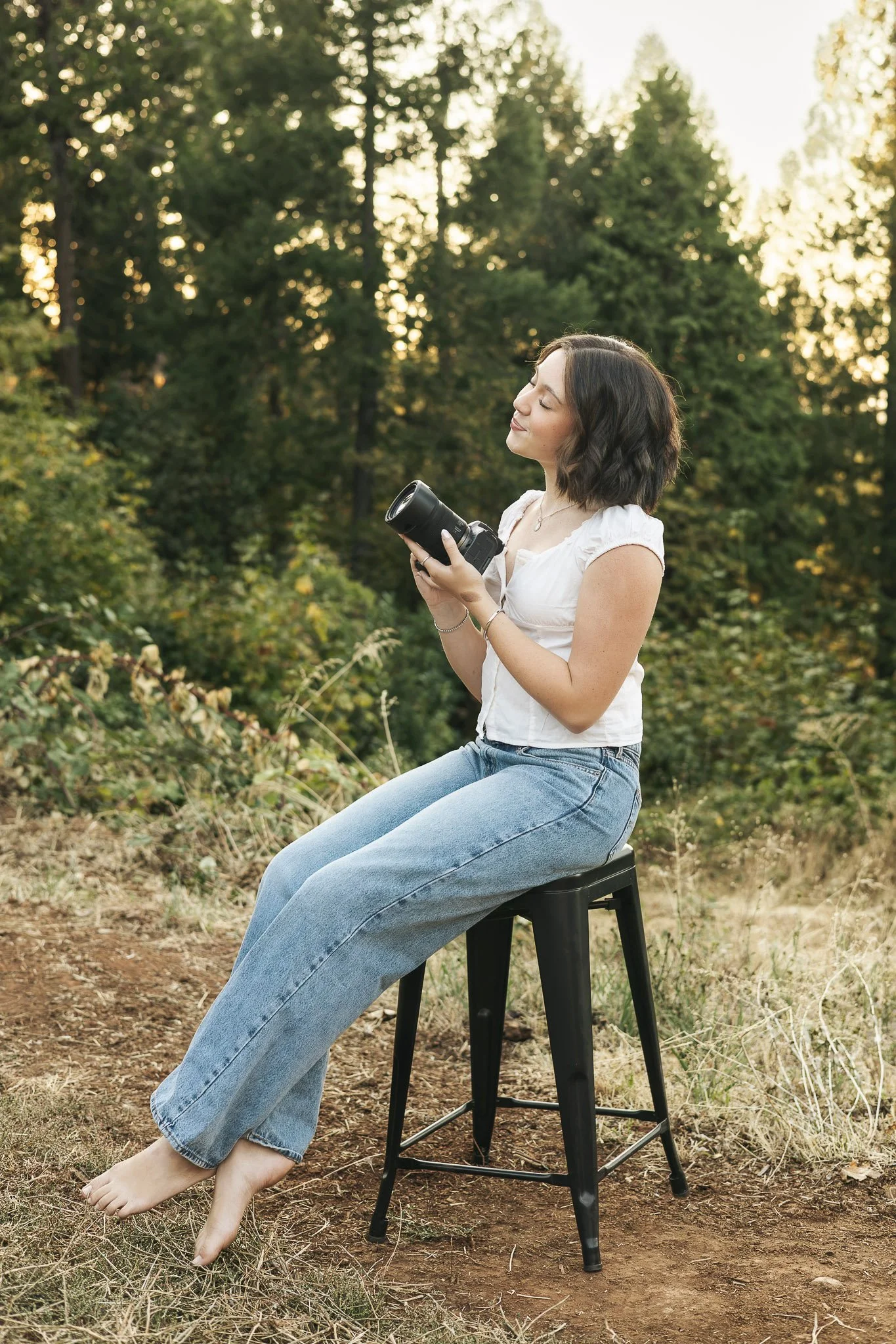 A woman sitting on a black stool outdoors in a natural setting, holding a camera, with her eyes closed and a serene expression, surrounded by trees and greenery during sunset.