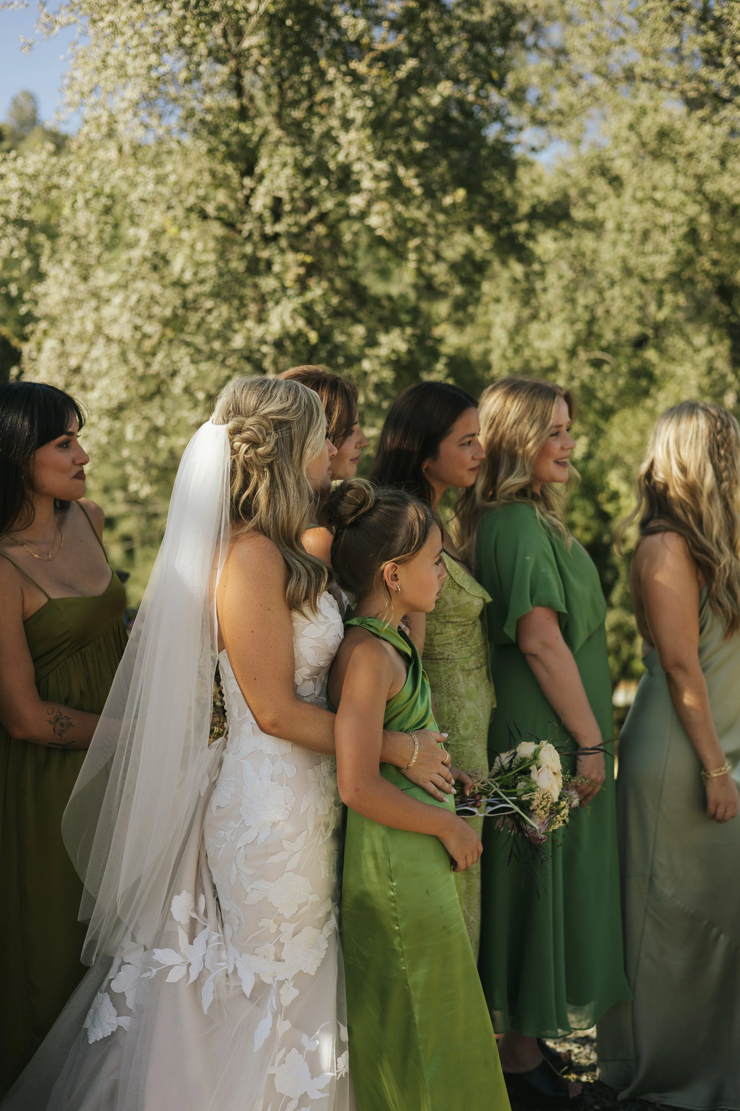 A group of women and girls standing outdoors during a wedding ceremony, with trees and greenery in the background. The bride is wearing a white lace dress with an embroidered floral pattern and a veil, while the other women are dressed in various shades of green and beige. Some are holding bouquets.