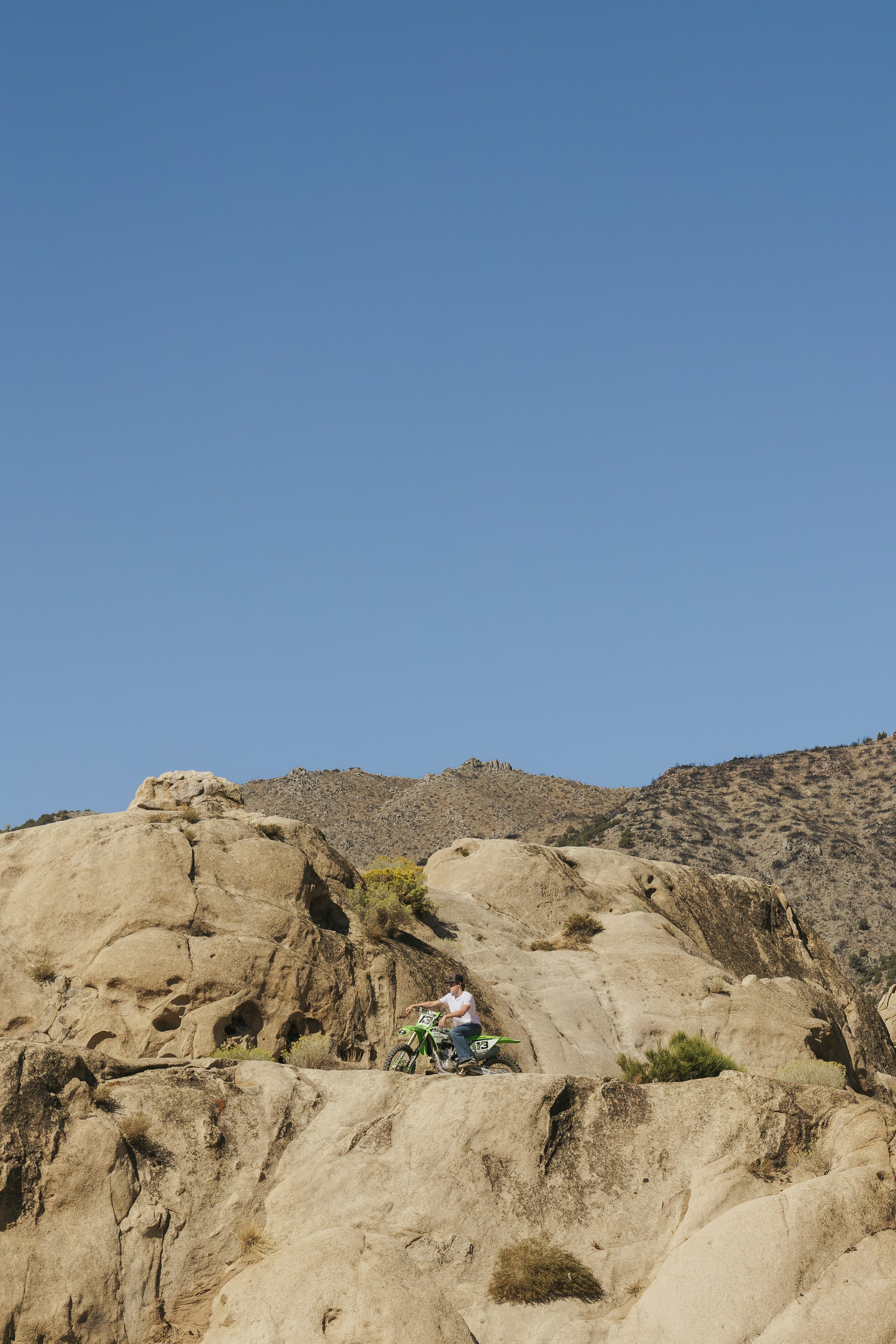 A person riding a green motorcycle with number 13 on a rocky, desert landscape with large boulders and mountains in the background under a clear blue sky.