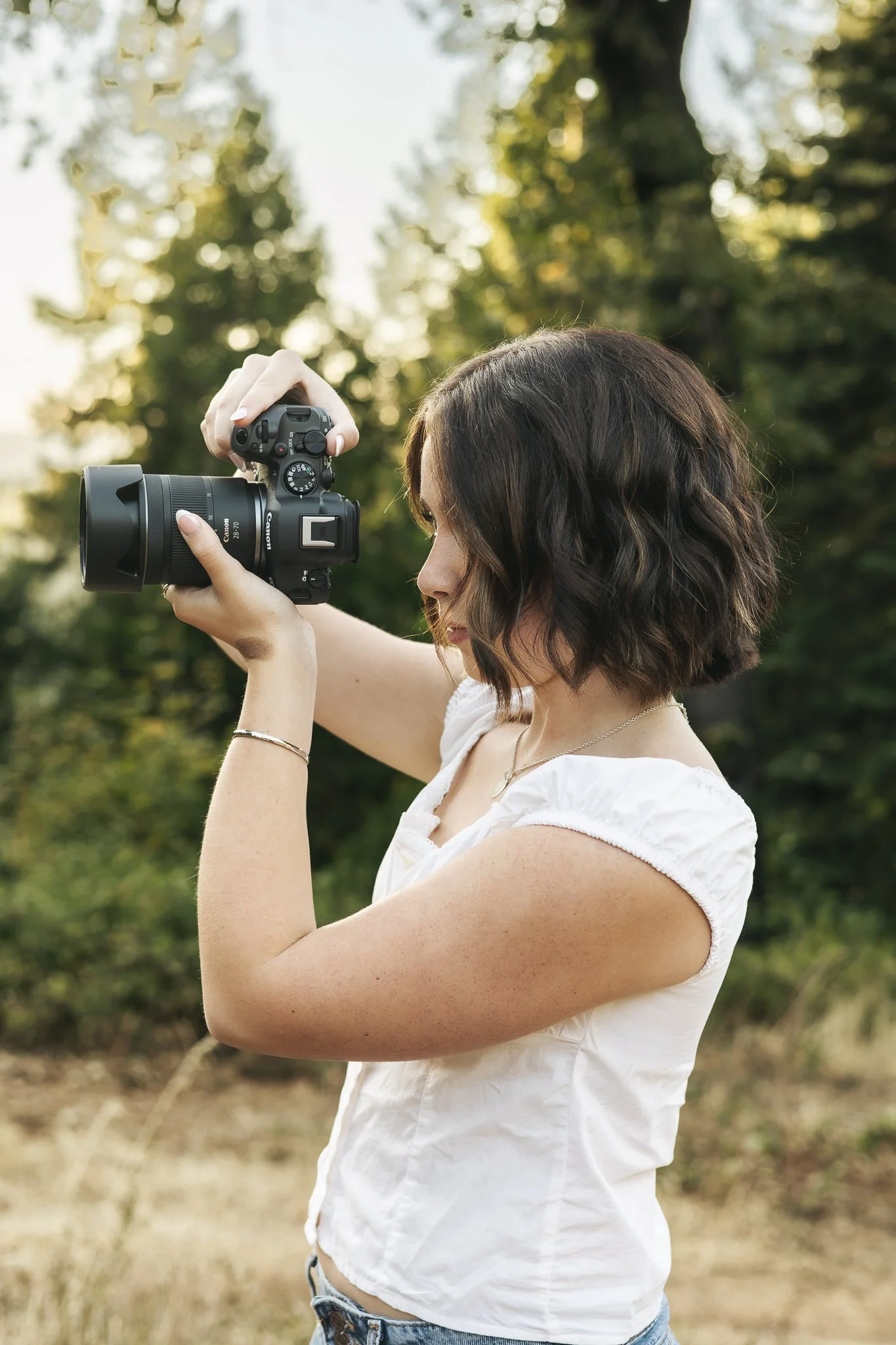 A woman with short, wavy brown hair wearing a white sleeveless top and denim shorts, standing outdoors holding a camera up to take a photo amidst trees and natural surroundings.