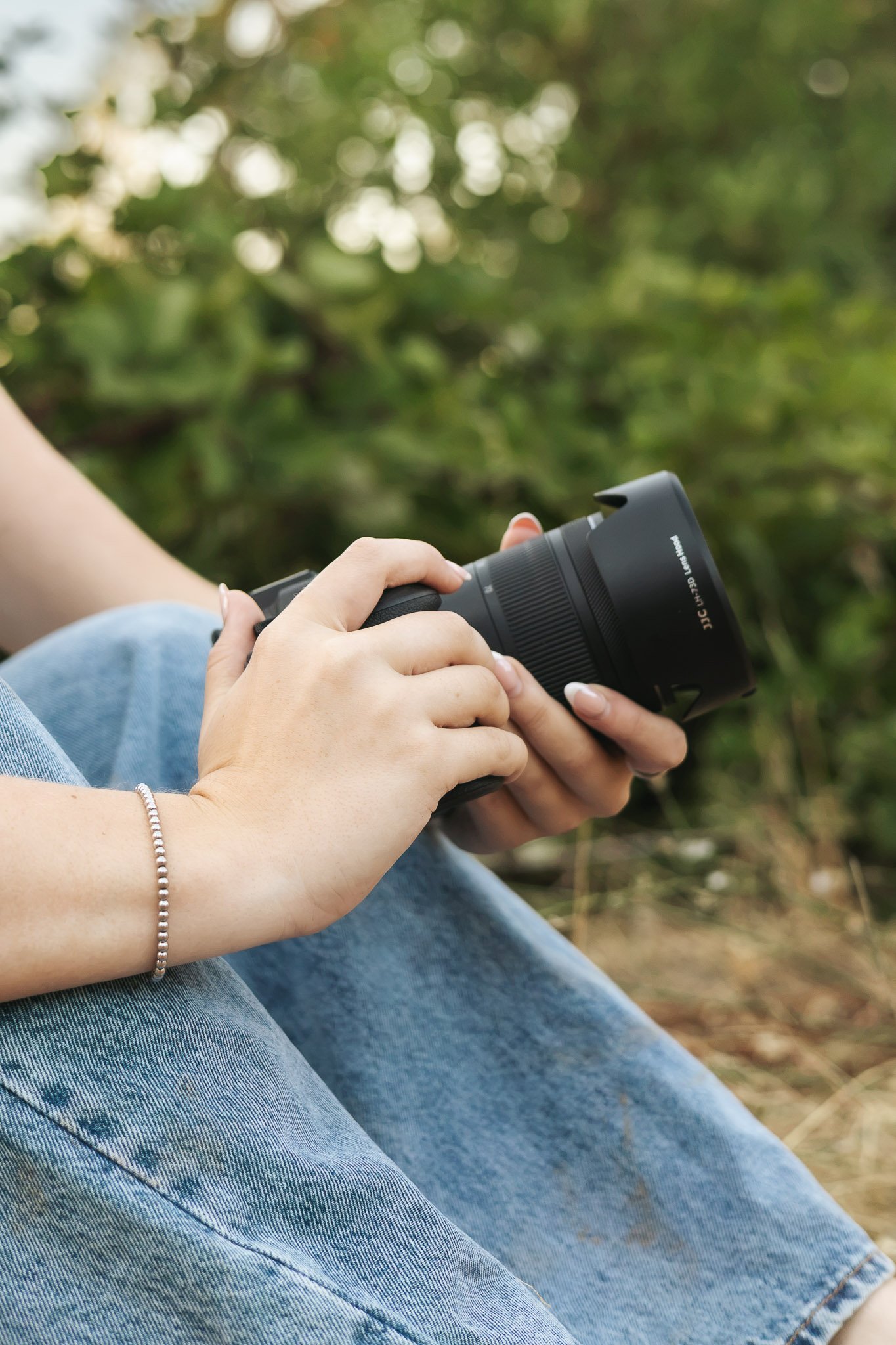 A person sitting outdoors holding a camera, with their legs crossed. The person is wearing blue jeans and a silver beaded bracelet.